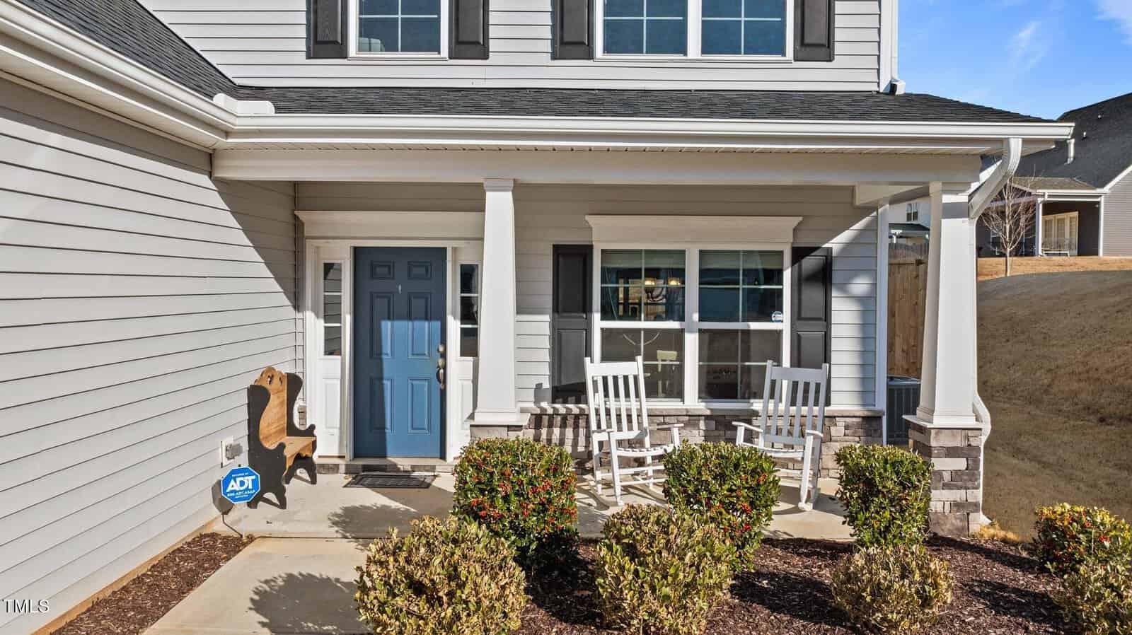 A front porch with two white rocking chairs, a blue front door, a wooden animal decoration, ADT security sign, white columns, shrubs along the walkway, and part of a two-story house in the background.