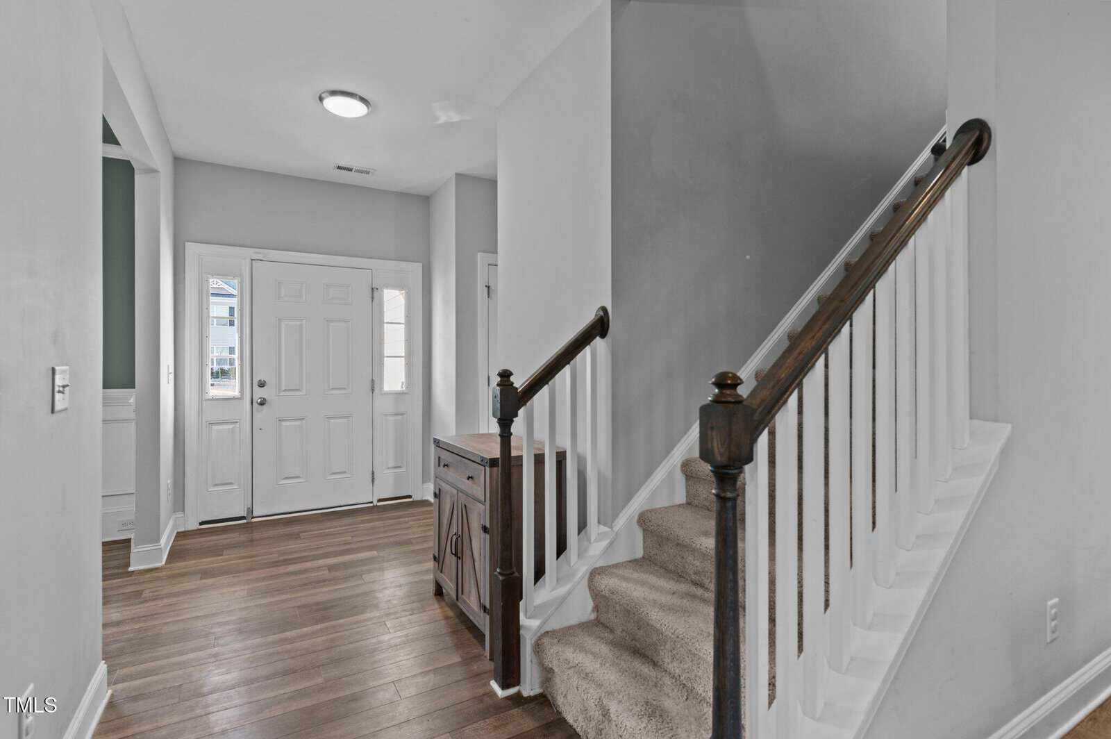 A residential entryway with hardwood floors, a white front door, and a carpeted staircase with a dark wooden handrail. A small wooden cabinet sits beside the stairs under a white wall.