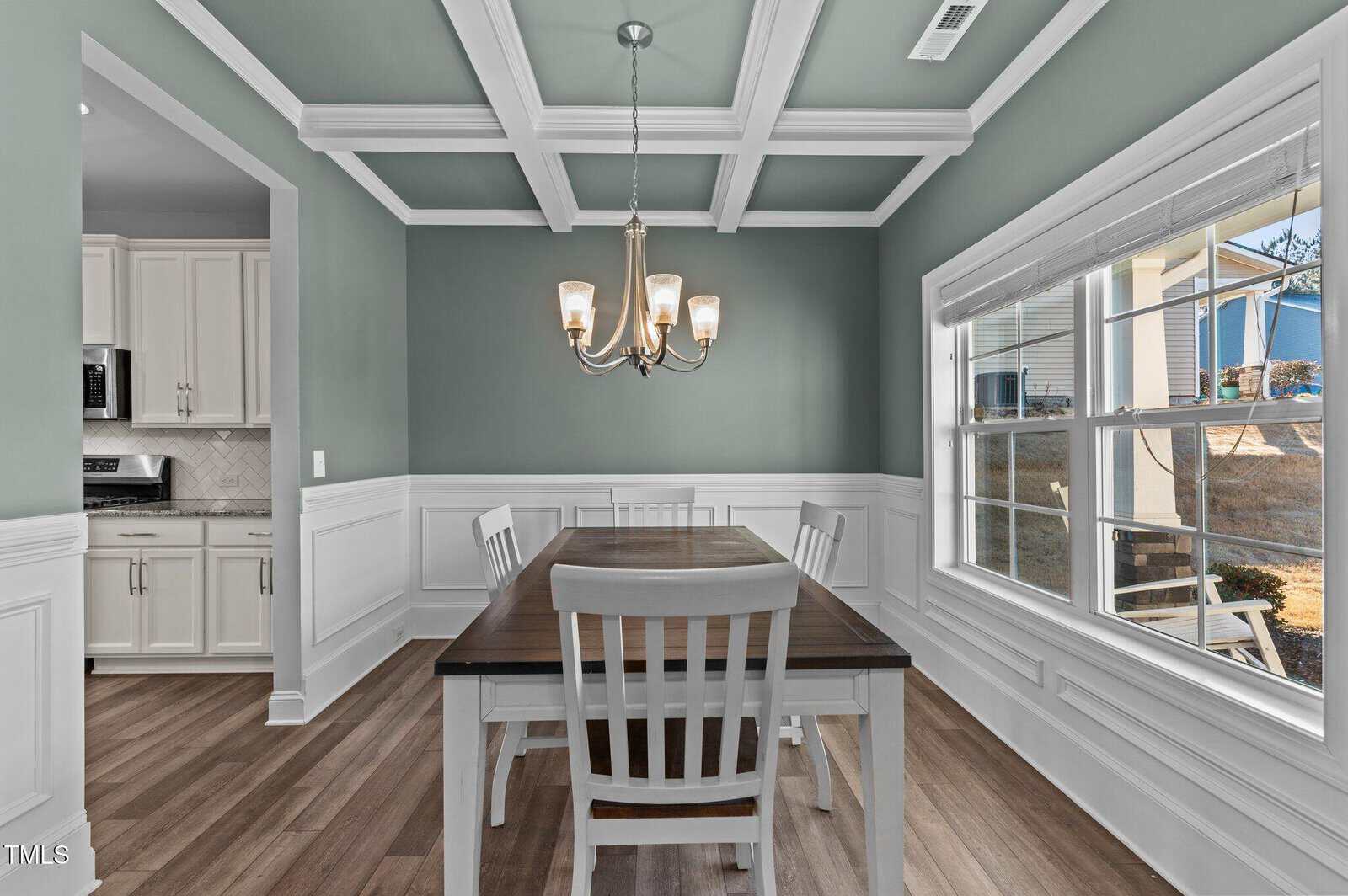 A dining room with a wooden table and four white chairs, a chandelier, wainscoting on the lower walls, coffered ceiling, wood flooring, and a large window next to the table; a kitchen is visible in the background.