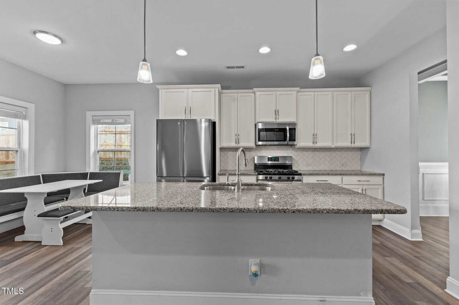 Modern kitchen with granite island, stainless steel appliances, white cabinets, pendant lights, a built-in bench dining area, and wood flooring. A double sink is on the island, and there is a herringbone backsplash behind the stove.