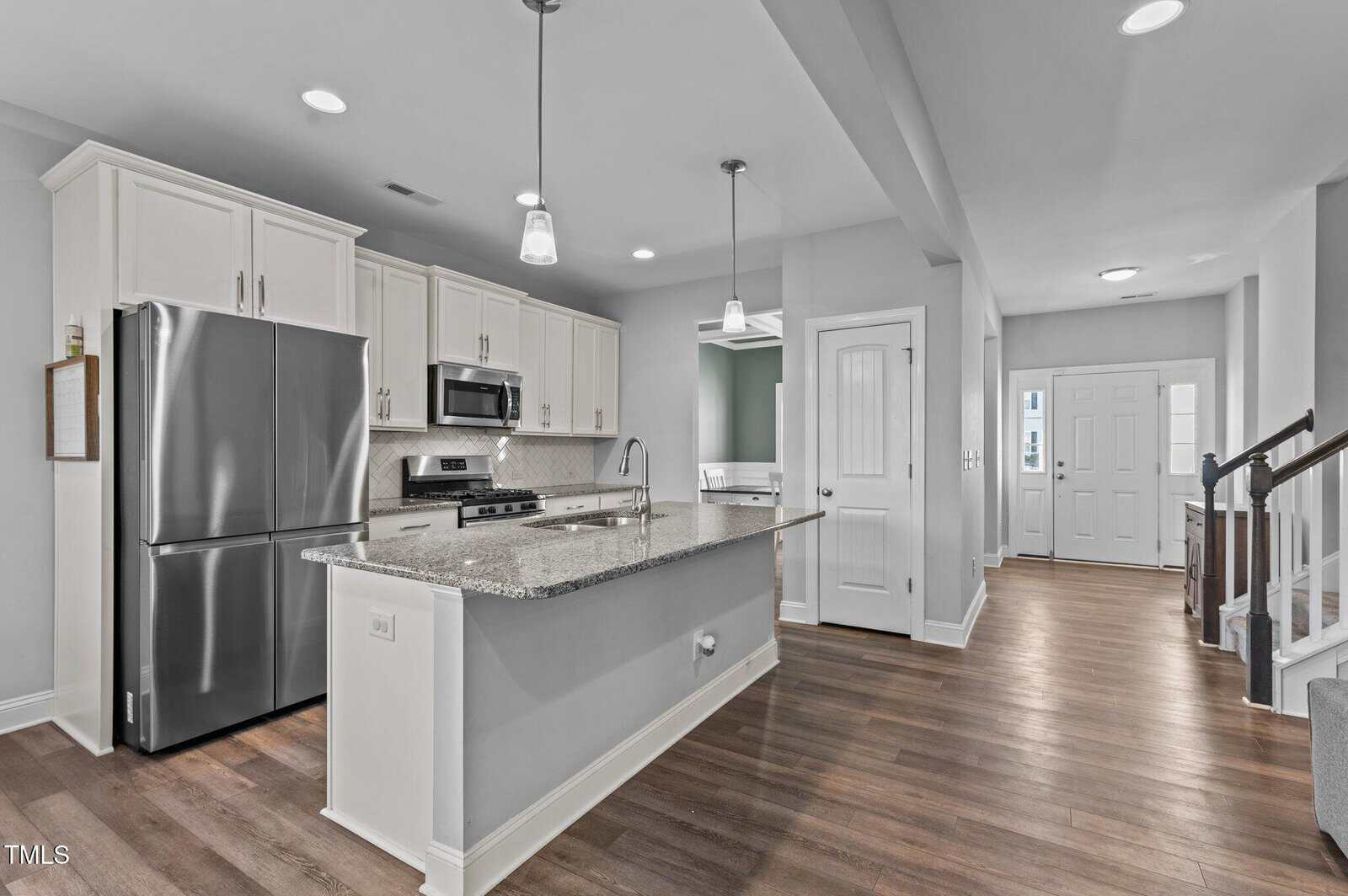 Modern kitchen with stainless steel appliances, white cabinets, granite countertop island, wood flooring, and pendant lights. Open layout leads to entryway and staircase. Walls are light gray with white trim.