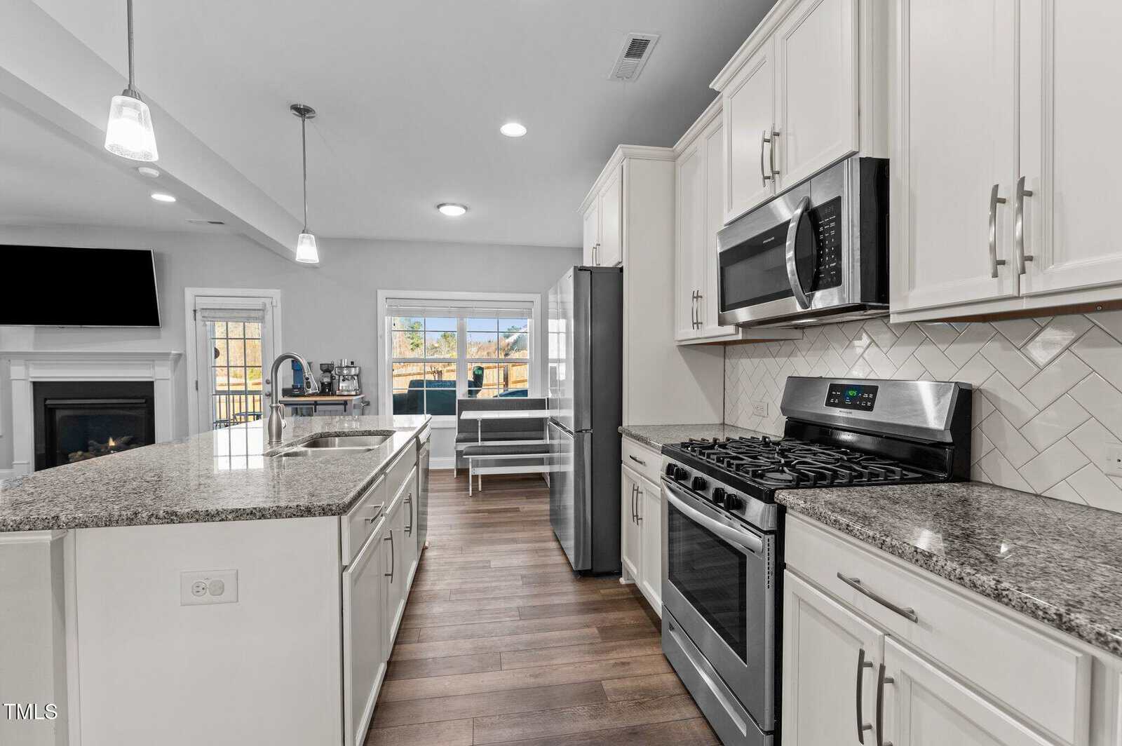 Modern kitchen with white cabinets, stainless steel appliances, gas stove, microwave, granite countertops, island with sink, and pendant lights. Open layout leads to a living area with fireplace and a view of the backyard through glass doors.