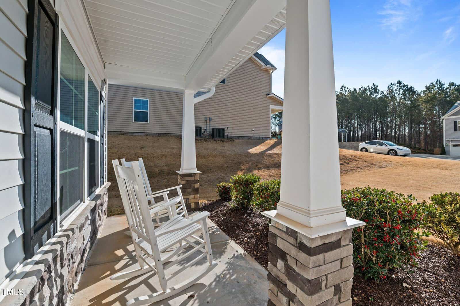 A covered front porch with two white rocking chairs, white columns with brick bases, a stone facade, trimmed bushes, and a view of neighboring houses and trees under a clear blue sky.