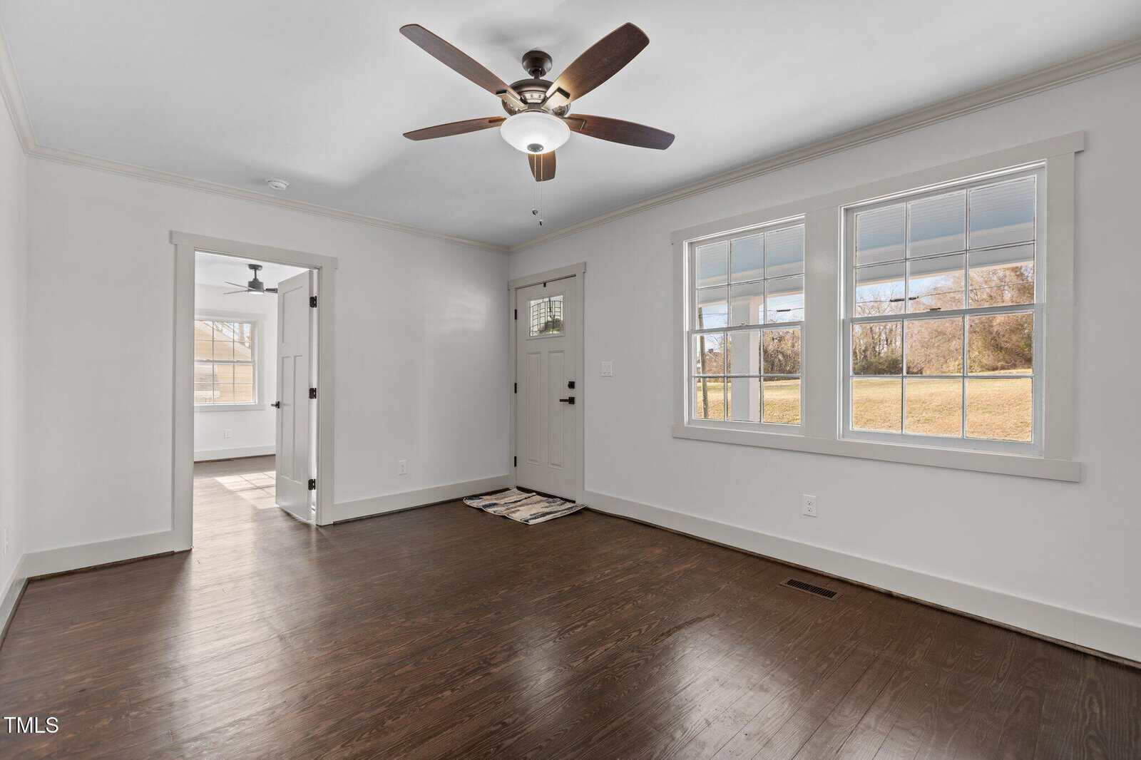 A room with dark hardwood floors, white walls, and a ceiling fan with a light. Three windows and a door lead outside. An open doorway reveals another room with similar flooring and a window.
