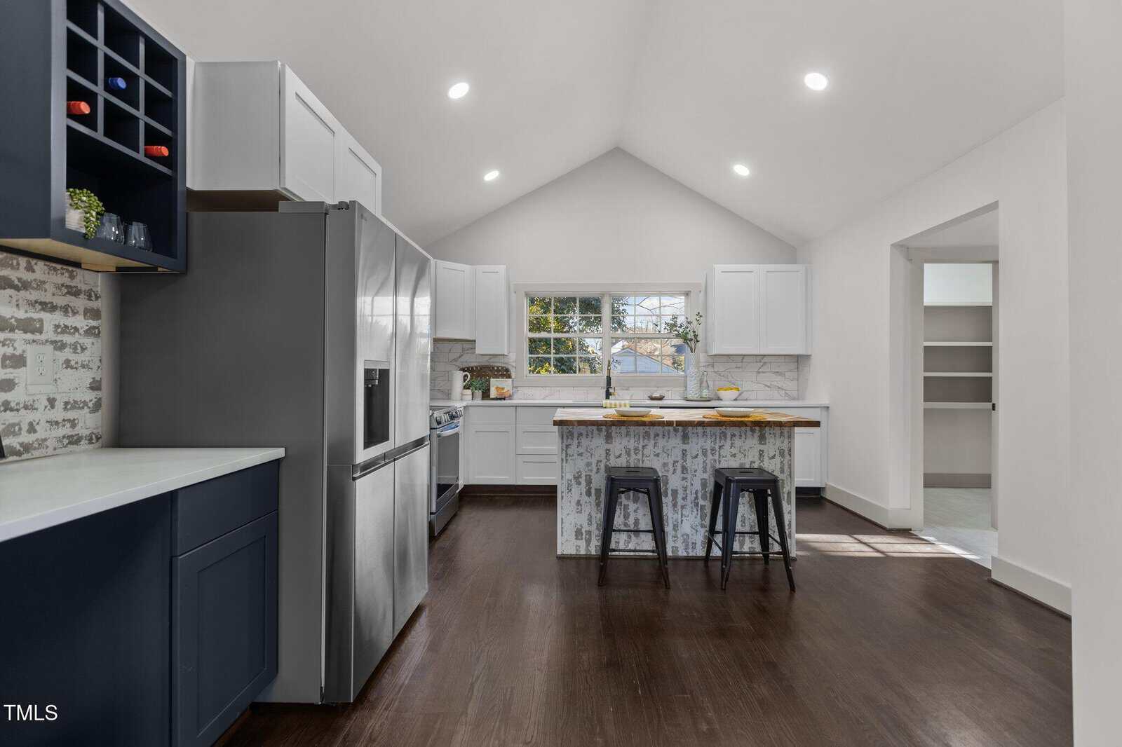 A modern kitchen with dark wood floors, white cabinets, a stainless steel refrigerator, an island with two black stools, and a brick accent wall. There is a window above the sink and open shelving to the left.
