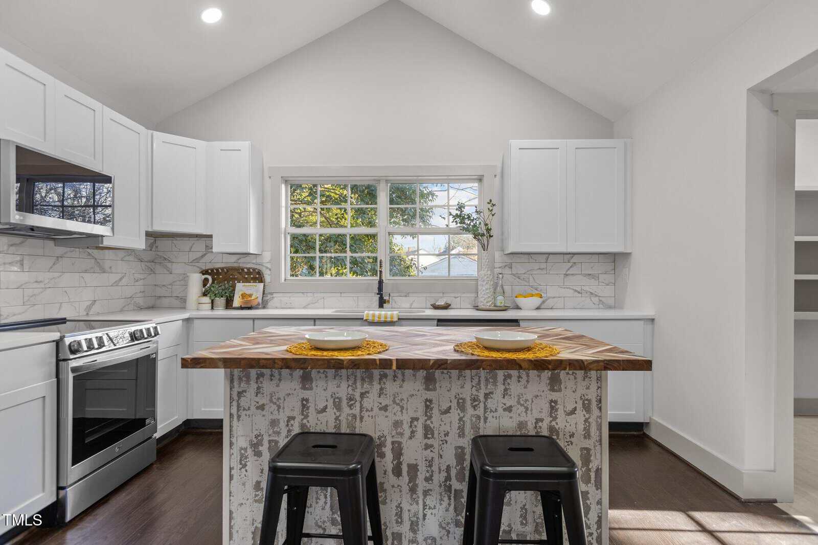 A modern kitchen with white cabinets, stainless steel appliances, a wood island with two black stools, place settings for two, and a large window letting in natural light. Marble backsplash and dark wood flooring.