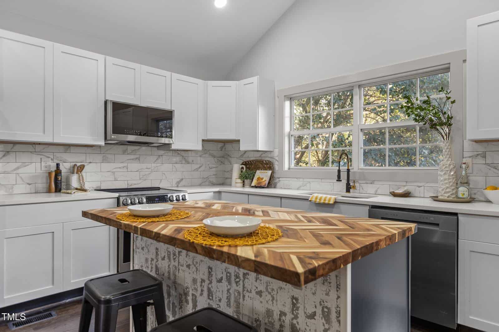 Modern kitchen with white cabinets, stainless steel appliances, a marble backsplash, and a large window. A wooden island with a patterned countertop and two stools has place settings with bowls and yellow placemats.
