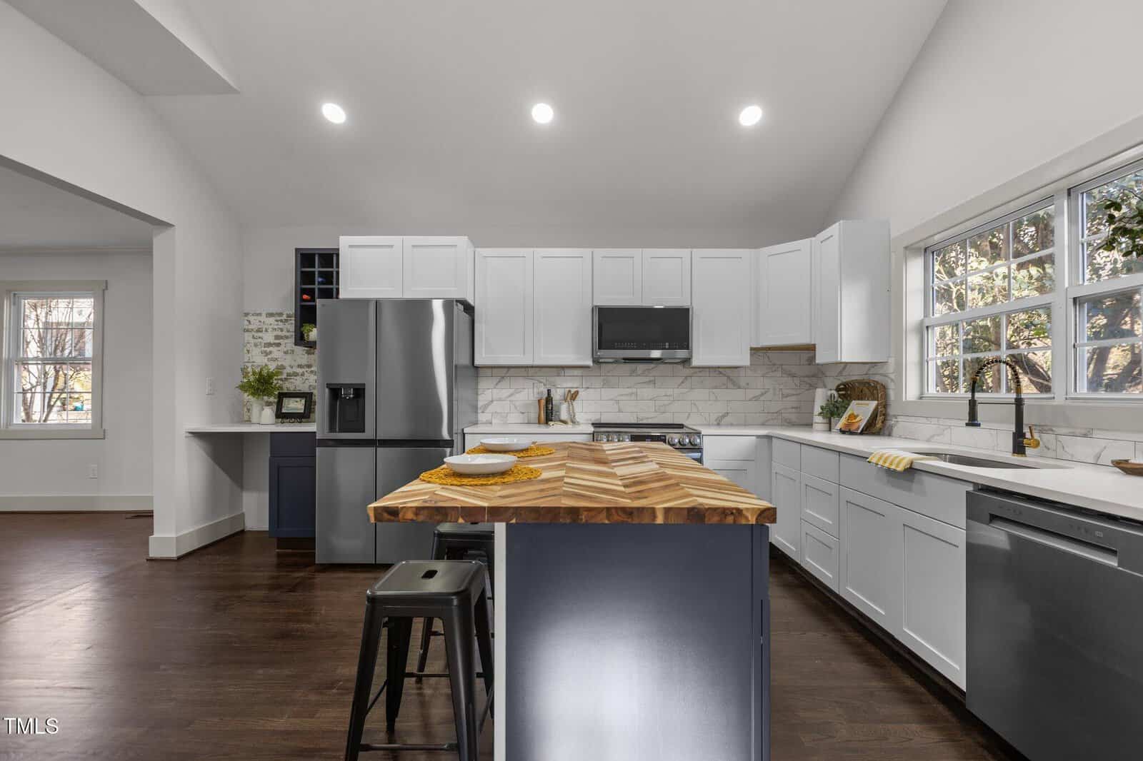 Modern kitchen with white cabinets, stainless steel appliances, a wood-patterned island with stools, dark wood floors, and large windows letting in natural light. Plates and cups are set on the island countertop.
