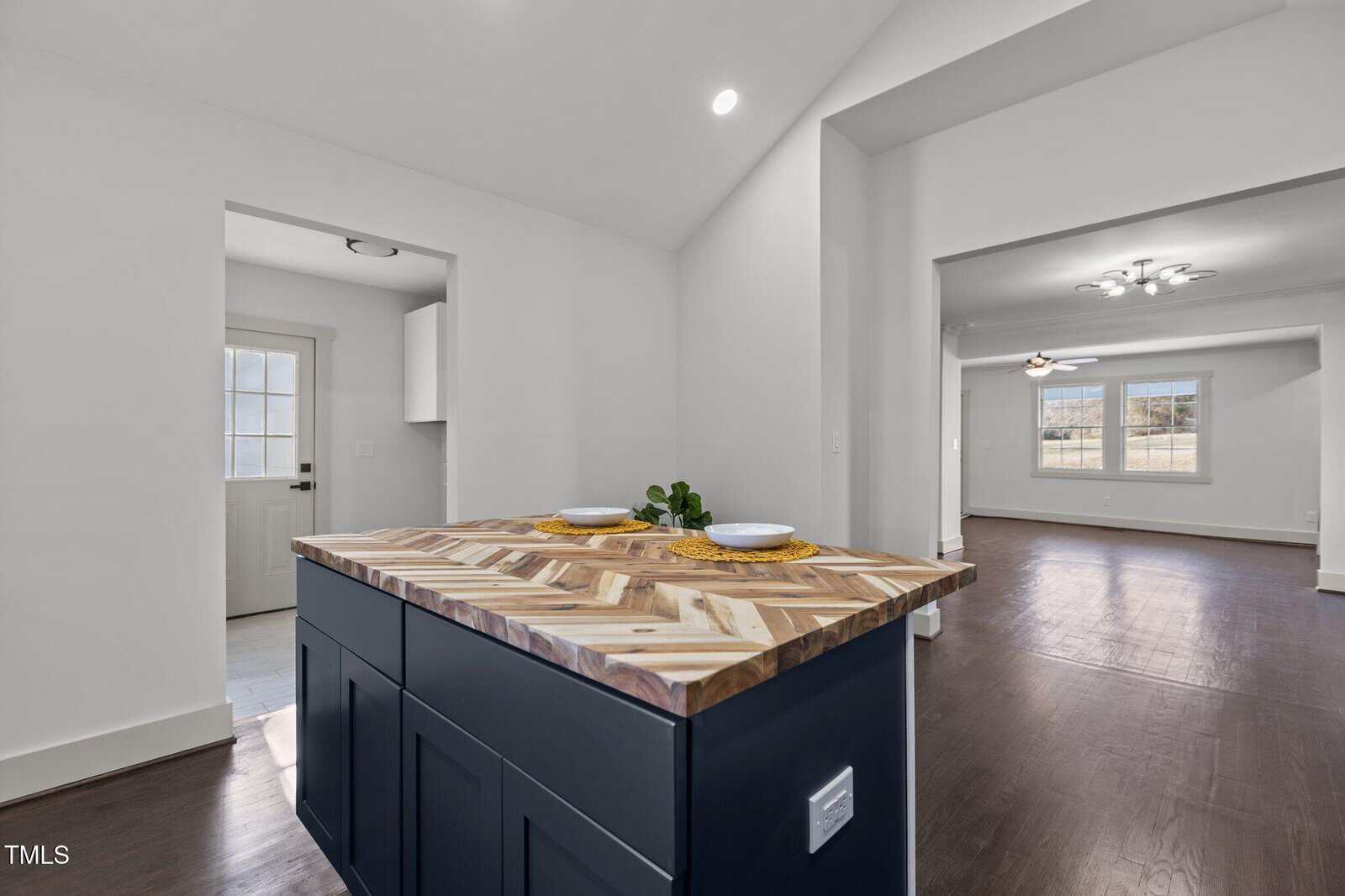 A modern kitchen island with a herringbone wood countertop, two plates, and a small plant. The space opens to a bright living room with dark wood floors and white walls. A door and window are visible in the background.