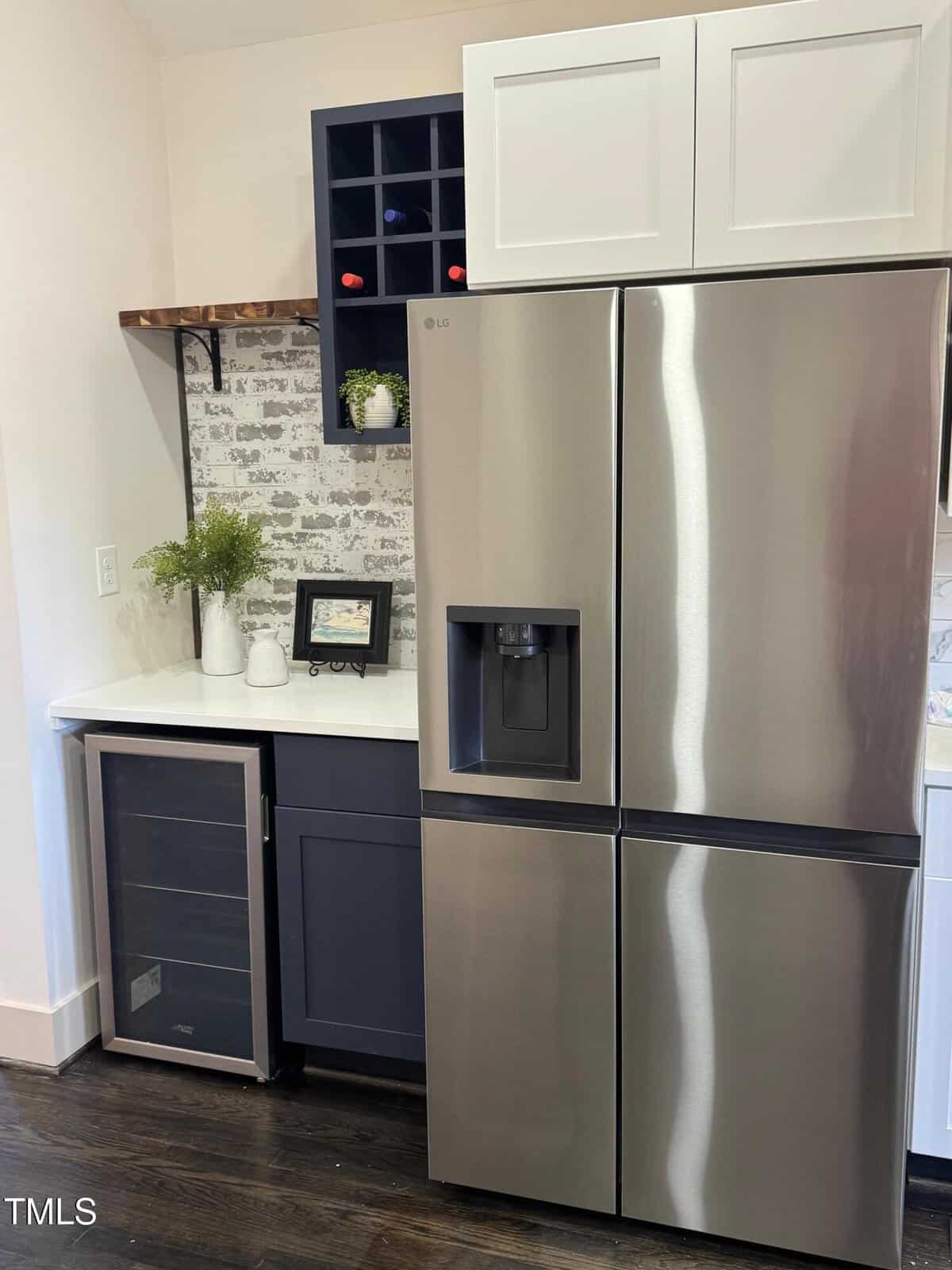 A modern kitchen corner with a stainless steel French door refrigerator, built-in wine cooler, navy blue cabinets, white countertop, potted plant, mugs, and a small wall wine rack holding four bottles.