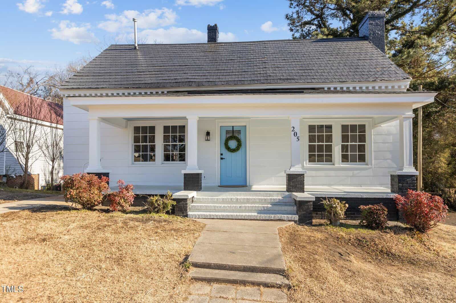 A white single-story house with a light blue front door, black shutters, and a wreath hanging on the door. There are bushes in front of the house, a concrete walkway, and a dry lawn. The house number 205 is visible.