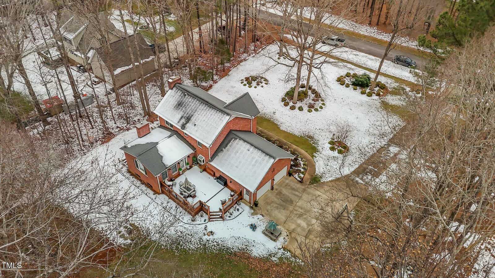 Aerial view of a red brick house partially covered in snow. The property features a large driveway, surrounding trees, and sparse snow on the ground. A road and neighboring houses are visible in the background.