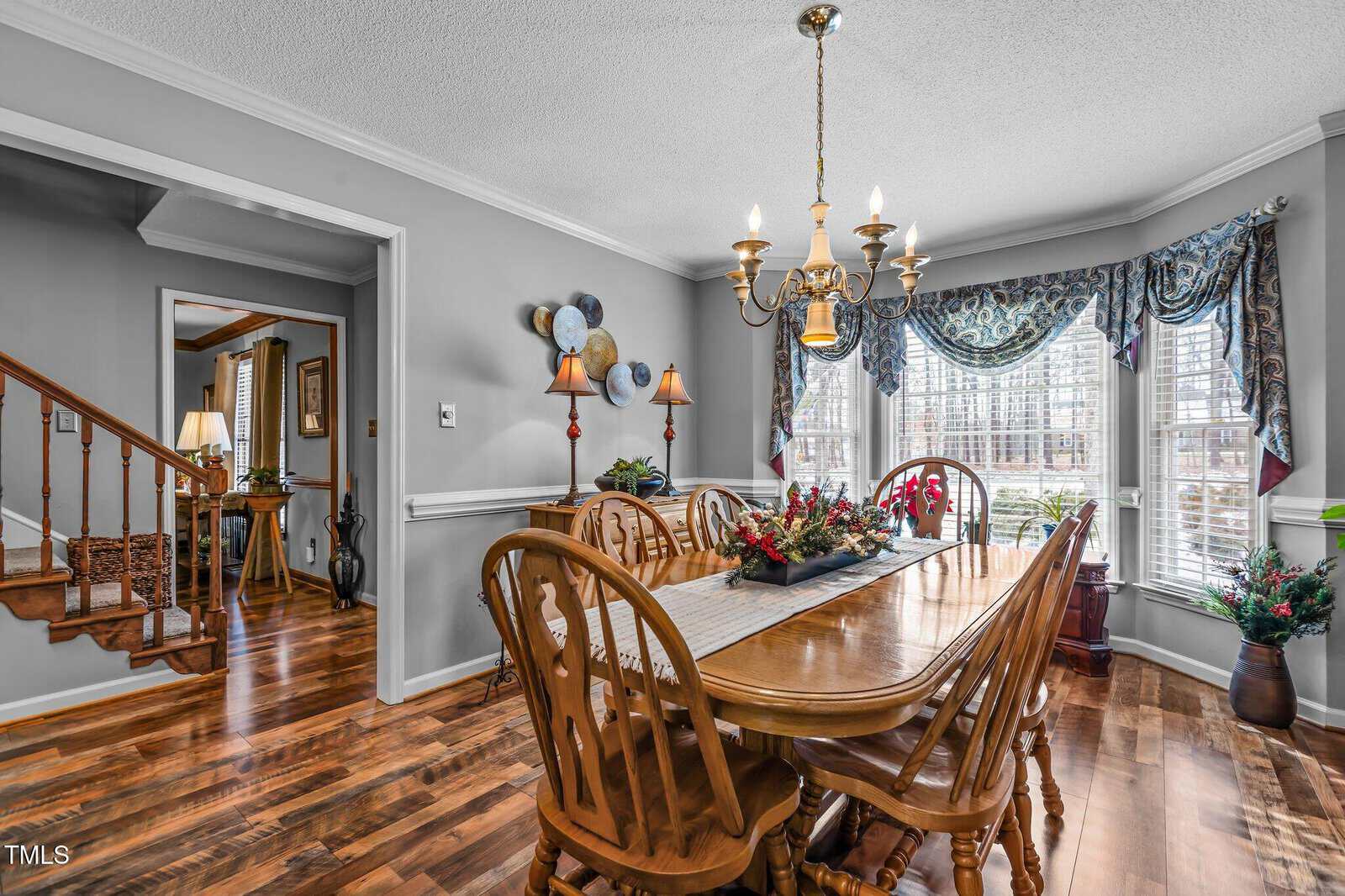 Dining room with a wooden table and six chairs. A chandelier hangs overhead. Large windows with patterned curtains allow natural light to enter. A plant and a decorative centerpiece are on the table. Walls are light gray with decorative plates.