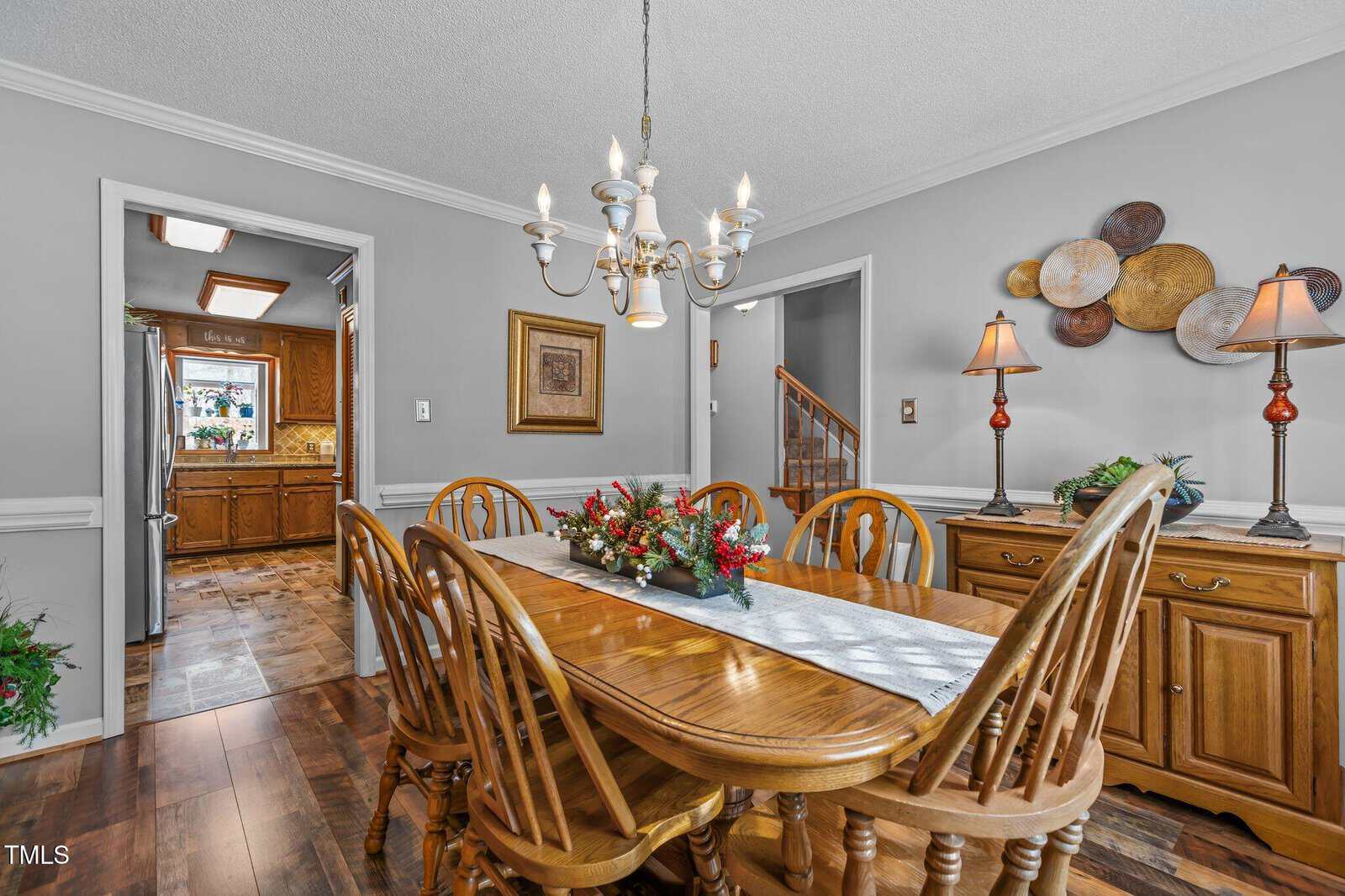 Dining room with a wooden table and eight chairs. A chandelier hangs above the table, which is decorated with floral arrangements. The room features a sideboard with lamps and a decorative wall piece. A kitchen is visible through an open doorway.