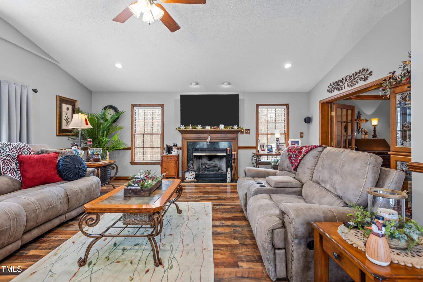 A living room with a vaulted ceiling, featuring two beige sofas and a wooden coffee table on a patterned rug. A fireplace with a TV above it is centered between two windows. The decor includes plants, pictures, and wall accents.