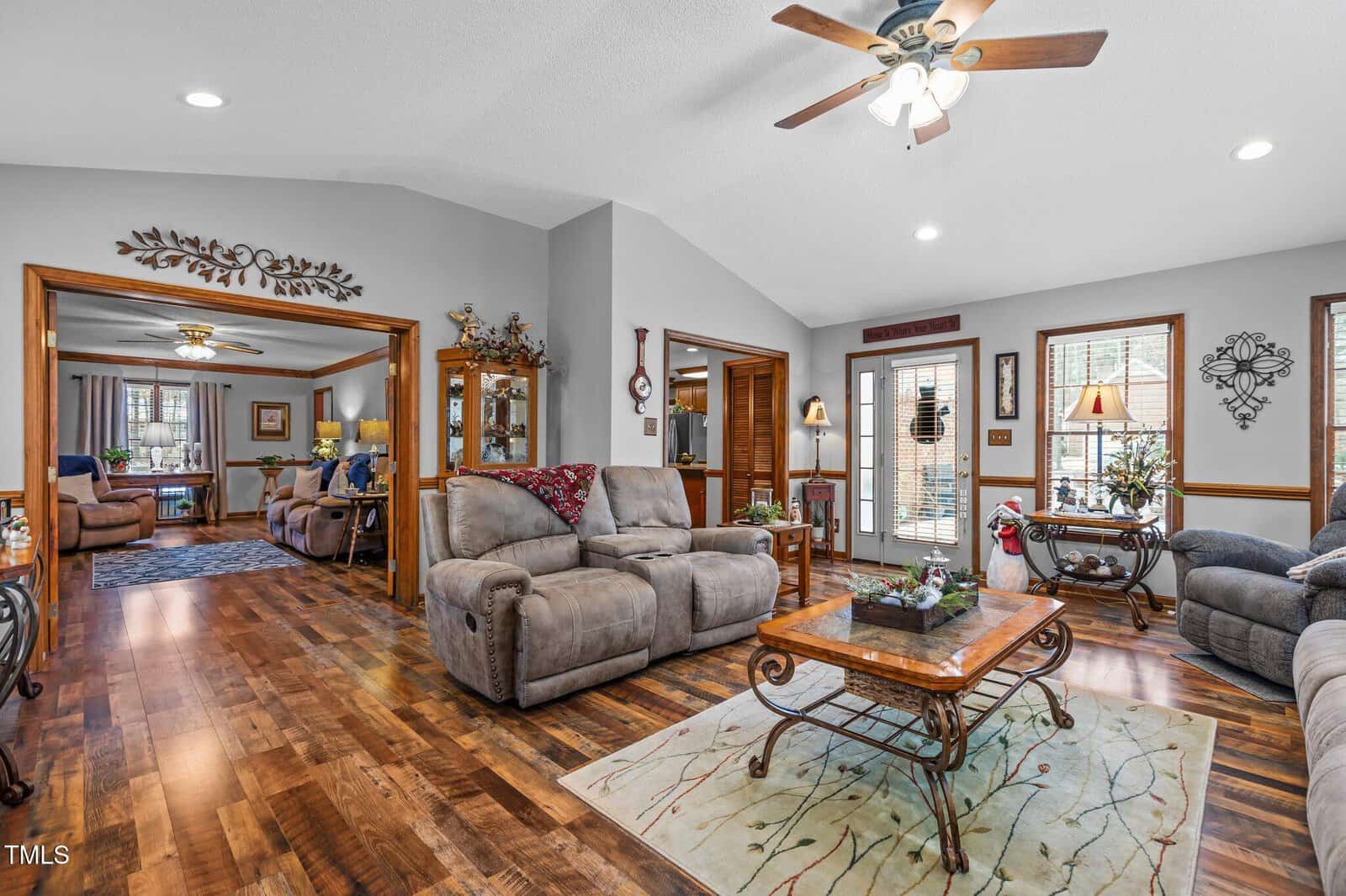 A cozy living room featuring two gray recliners and a sofa. A wooden coffee table sits on a patterned rug. Ceiling fan with lights above. French doors and windows provide natural light. An adjacent room with more seating is visible in the background.