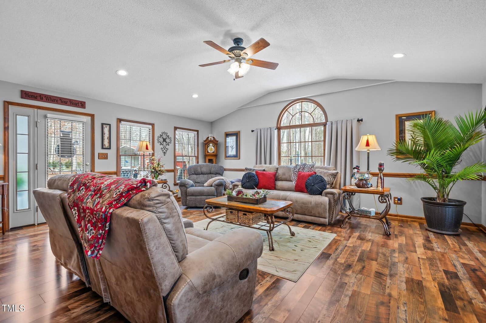 A spacious living room with hardwood floors, beige sofas, and a wooden coffee table. There are large arched windows with curtains, ceiling fan, and a tall potted plant. Decor includes a grandfather clock and wall signs.