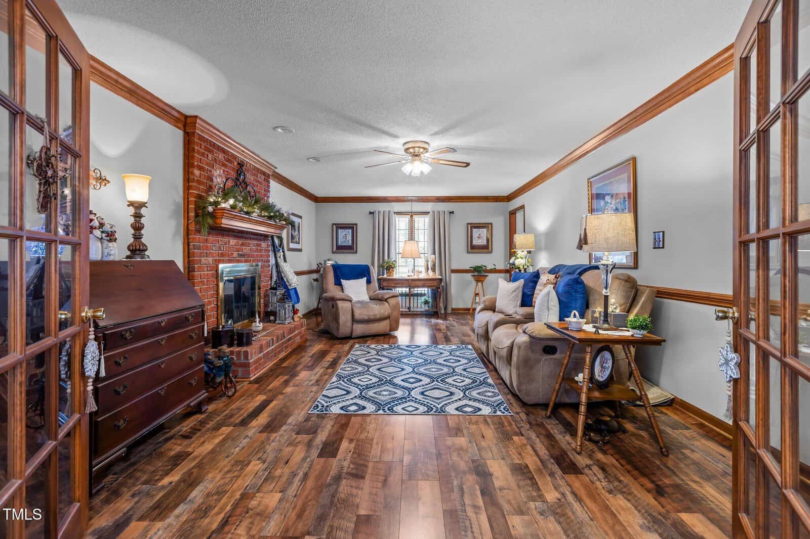 A cozy living room with wooden floors and brick fireplace, adorned with greenery. The room features a plush sofa, armchair, and side tables with lamps. A patterned rug and ceiling fan add comfort and style, with a window view at the far end.