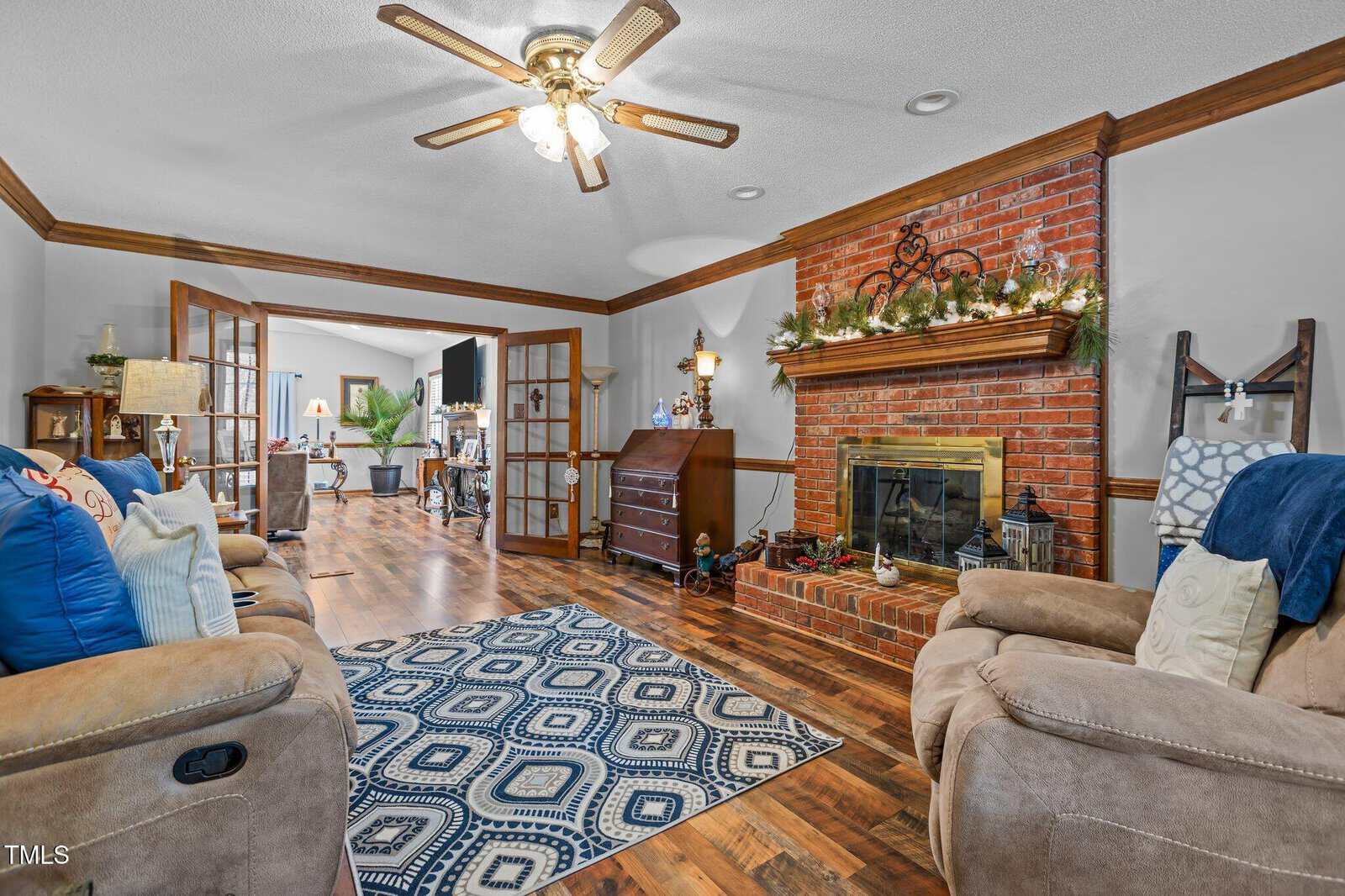 A cozy living room with a brown brick fireplace adorned with greenery and lights. Two beige recliners face a blue and white patterned rug. Wooden floors and trim create a warm ambiance. French doors open to another room with visible decor.