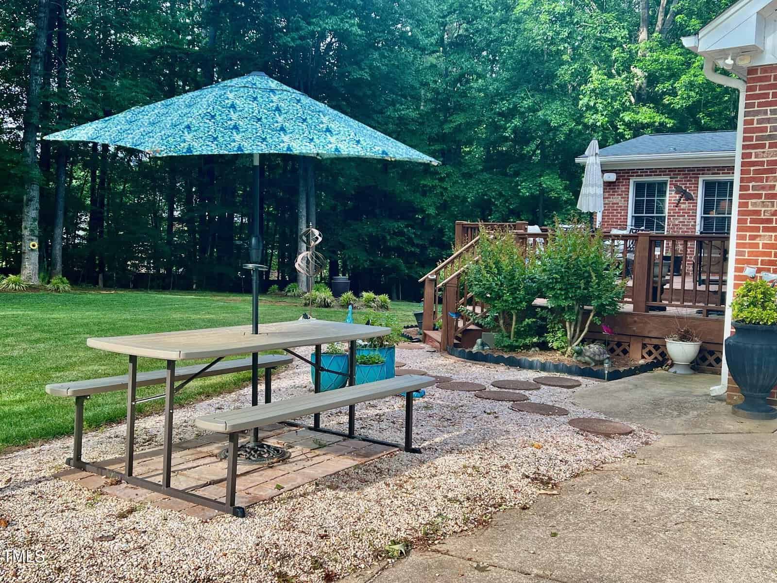 A picnic table with attached benches sits on a gravel area under a blue patterned umbrella, next to a grassy yard and brick house with a wooden deck surrounded by trees and garden plants.