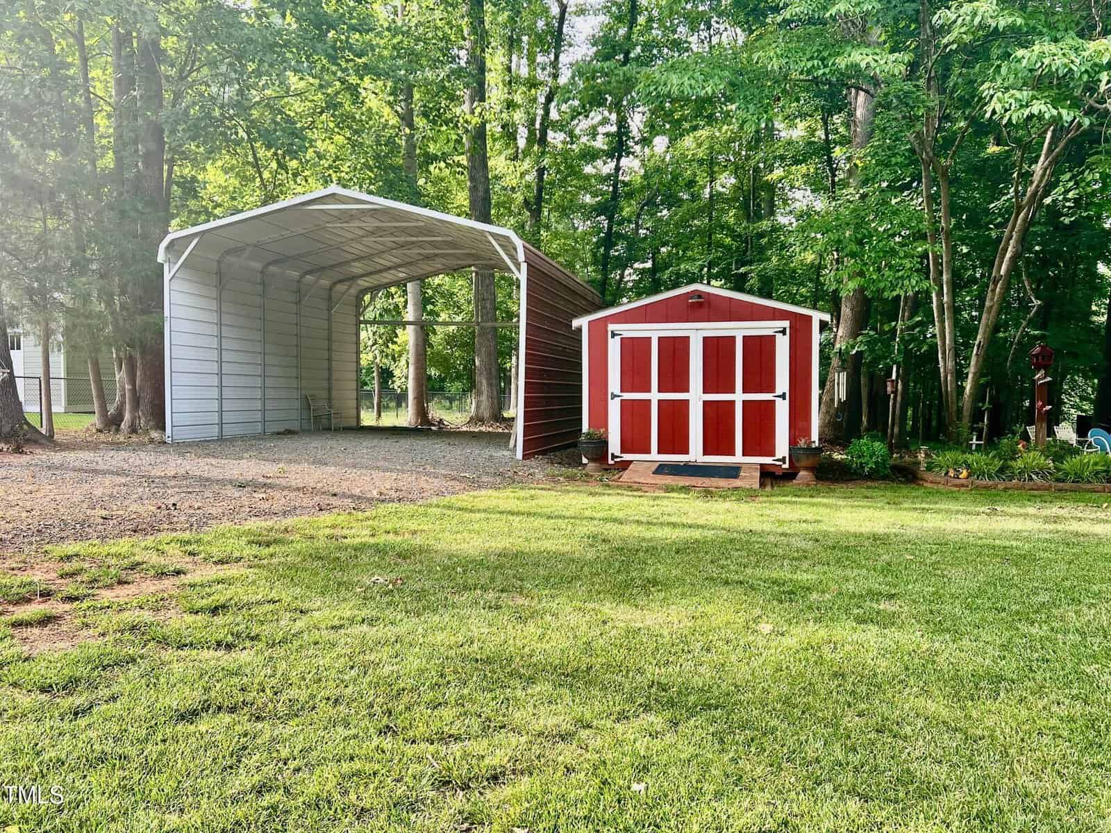A metal carport with an open front stands next to a red and white wooden shed. Both are situated on a grassy lawn bordered by trees in the background.