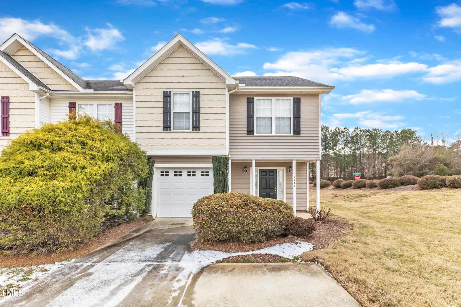 Two-story beige townhouse with black shutters and a single-car garage. Bushes line the front and side, and a driveway leads to the entrance. Grass and shrubs border an open field under a partly cloudy sky.