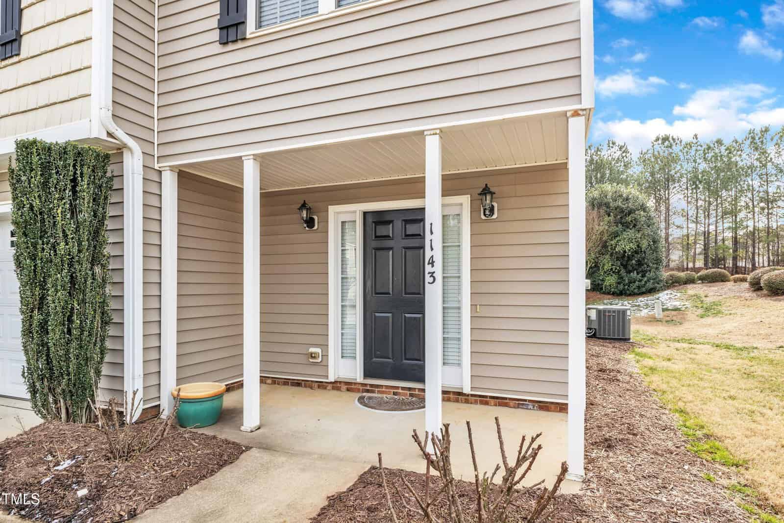 A beige house with vertical and horizontal siding, a black front door, white pillars, a welcome mat, two wall-mounted lights, and a small concrete porch. Mulched landscaping and a potted plant are in front. Trees and HVAC units are visible to the right.