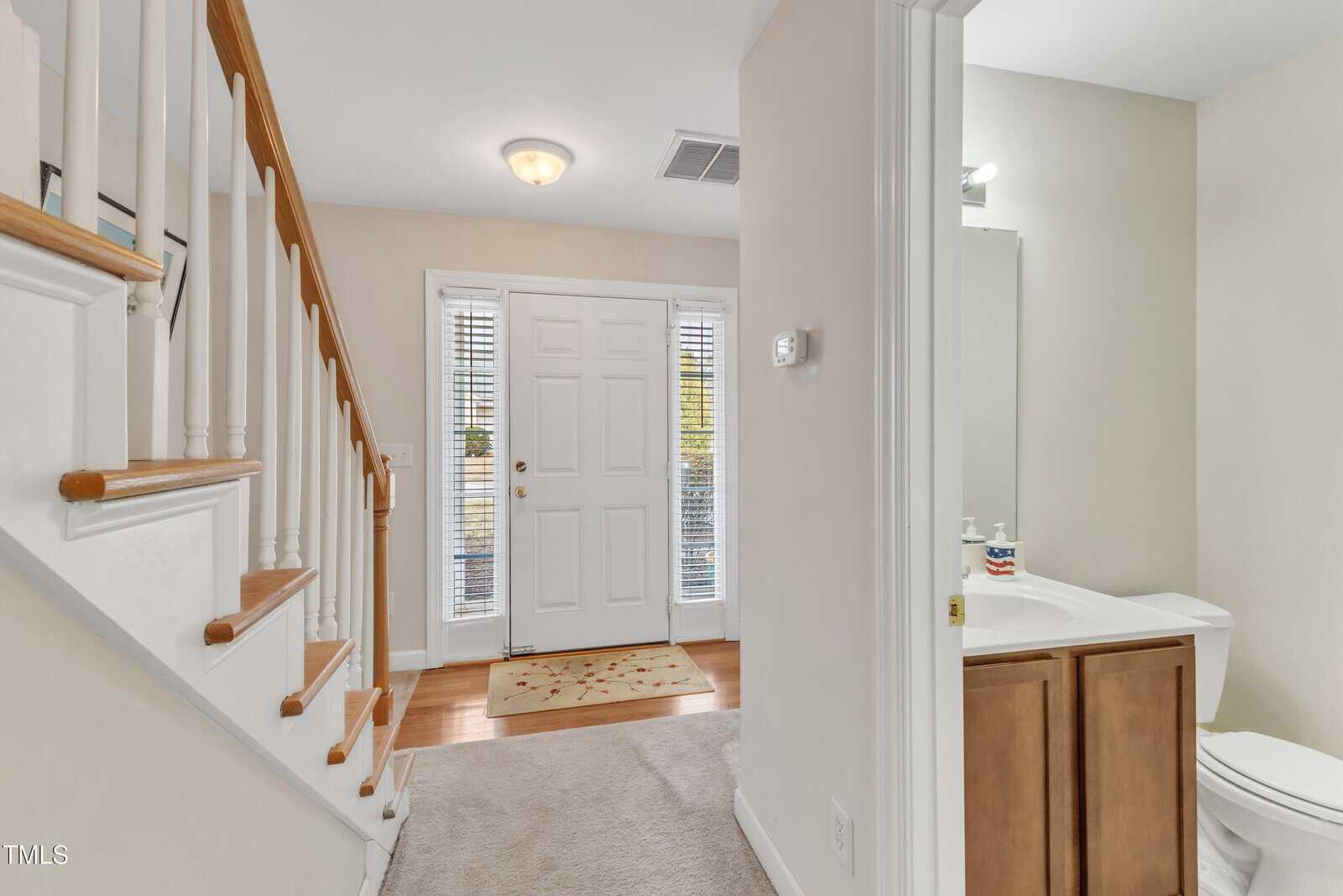 A view of a home's entryway with a staircase on the left, a white front door straight ahead, and a bathroom with a sink and toilet visible on the right. The area is well-lit and has neutral-colored walls.