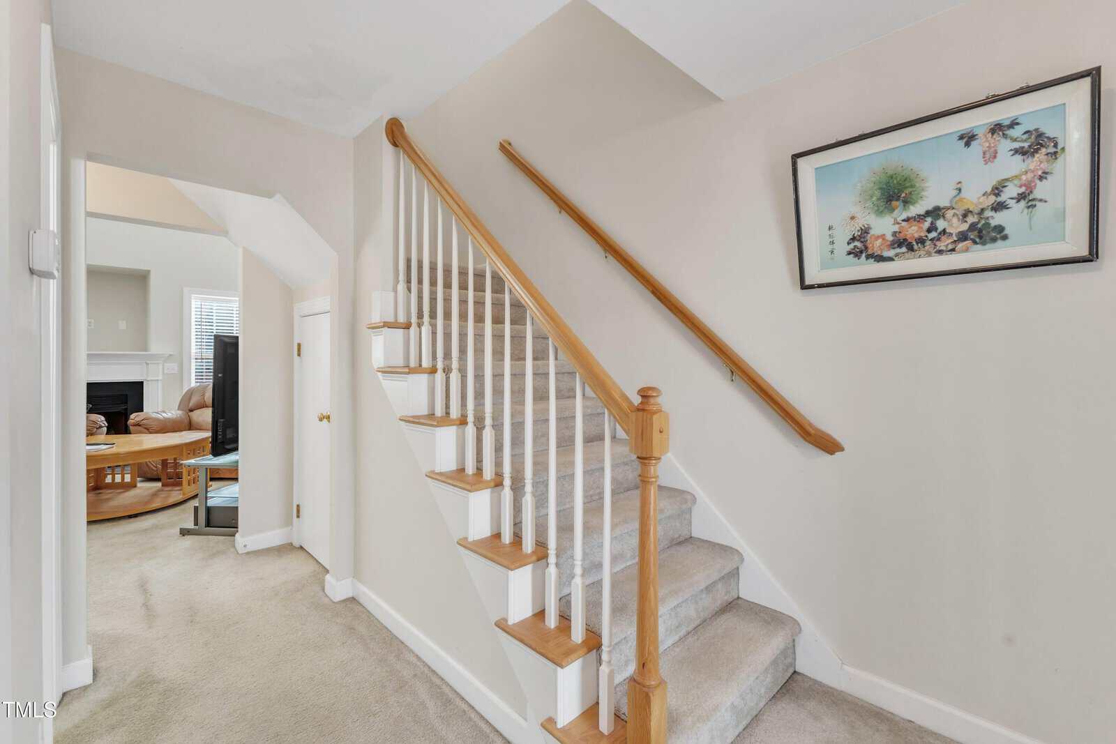 A carpeted staircase with a wooden handrail is on the right side of a hallway. A framed floral print hangs above the stairs, and a small door is below the staircase. An open doorway leads to a living room.