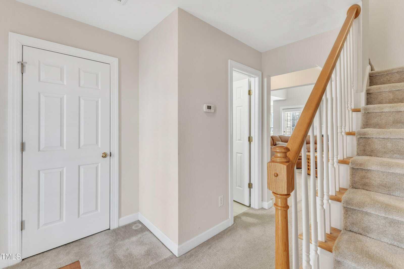 A beige-carpeted hallway with a white door on the left, a thermostat on the wall, an open doorway leading to a bright room, and a carpeted staircase with a wooden handrail on the right.