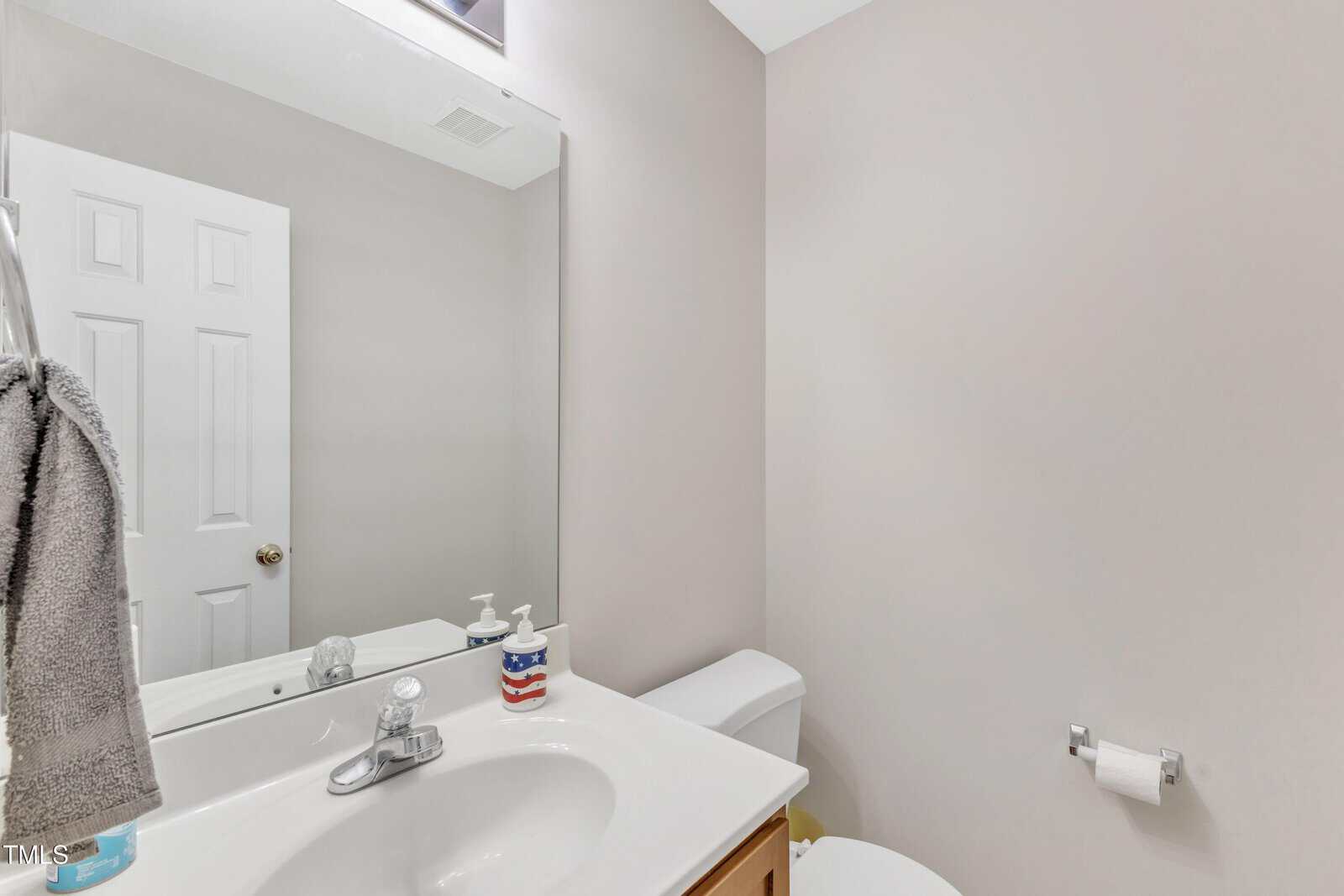 A small bathroom with a white sink, a mirror, a towel, soap dispensers, a toilet, and a toilet paper roll on a beige wall.