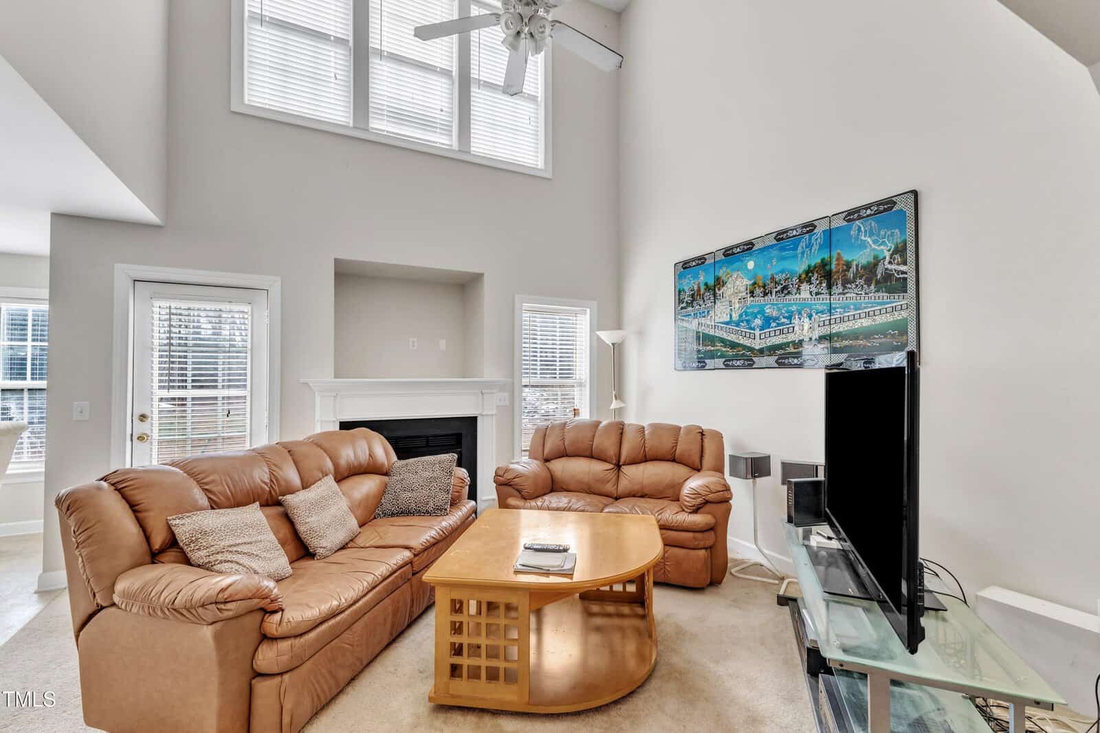 A bright living room with two tan leather sofas, a wooden coffee table, a TV on a glass stand, a fireplace, large windows, and a colorful cityscape painting on the wall.