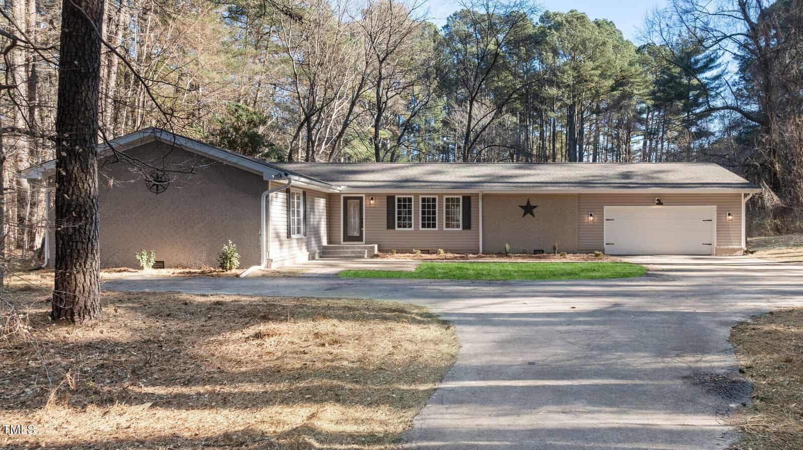 Single-story house with a gray exterior, attached two-car garage, and a front porch. The house has white-trim windows and a star decoration on the front wall. Surrounded by tall trees and a curved driveway.