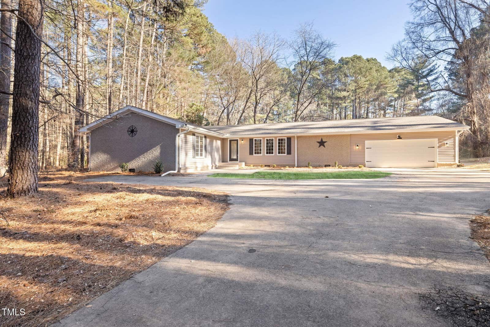 Single-story gray house with a two-car garage, front porch, and large driveway set among tall trees. The yard is mostly pine straw with woodland surroundings and a clear sky overhead.