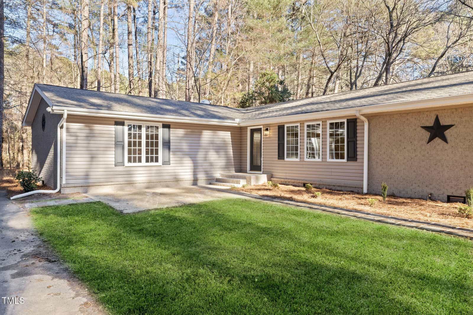 Single-story house with beige siding and multiple large windows, set against a backdrop of tall trees. The front yard has green grass and a concrete walkway. A black star decorates the exterior wall near the entrance.