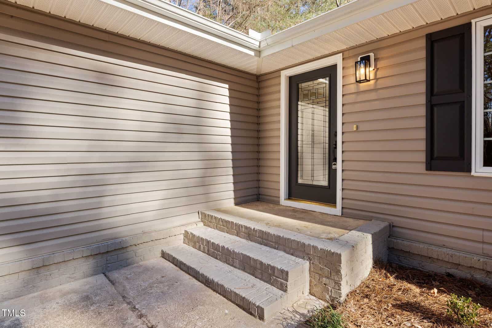 Beige vinyl-sided house exterior with a black-framed glass door, steps made of brick leading to the entrance, a wall-mounted light, and a small concrete porch area.