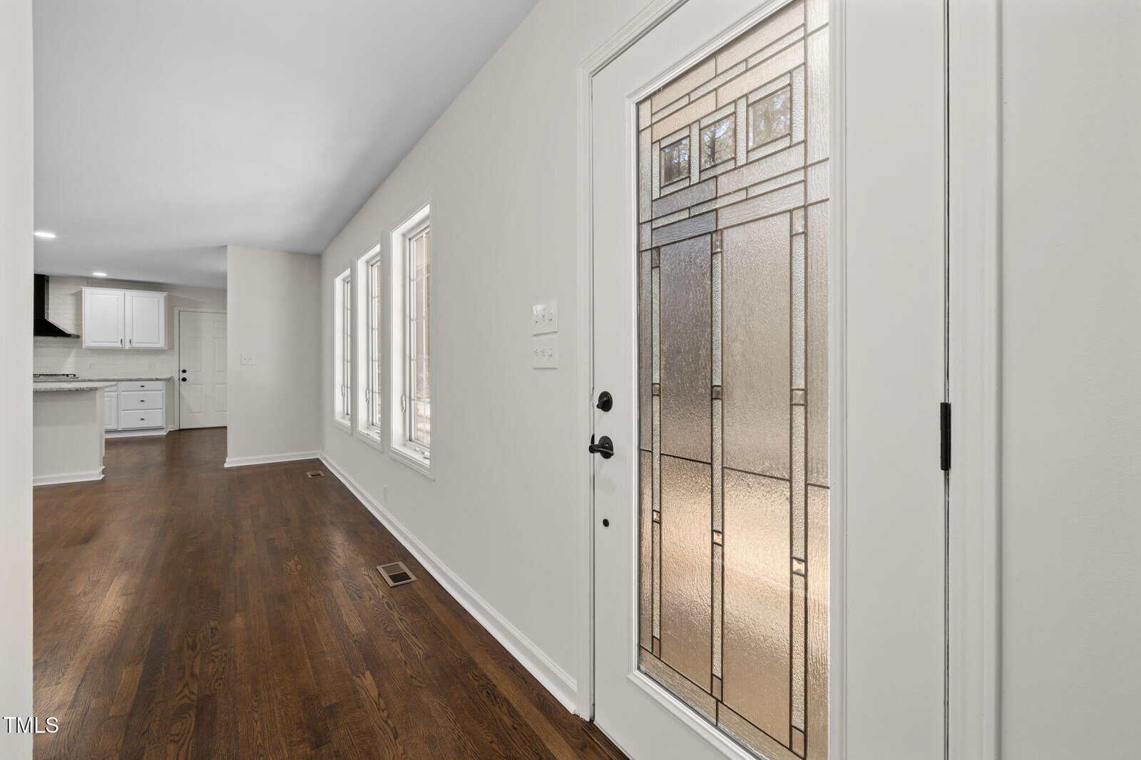 A hallway with hardwood floors, three windows on the left, and a glass-paneled front door on the right. The hallway leads to a white kitchen visible in the background.
