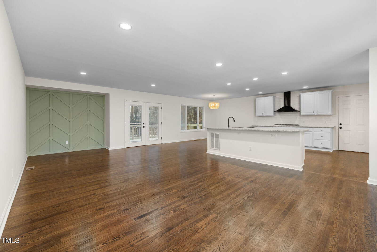 Open-concept living area with hardwood floors, white walls, recessed lighting, green accent wall, large windows, and a kitchen featuring white cabinets, a center island, and a black range hood.