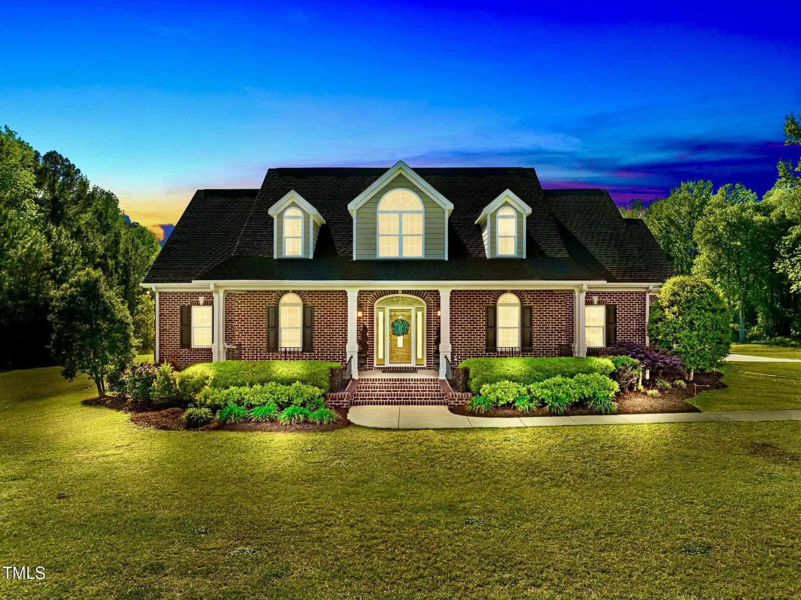 A two-story brick house with three dormer windows, a covered front porch, and exterior lights on, surrounded by manicured shrubs and lawn, set against a twilight sky with trees in the background.