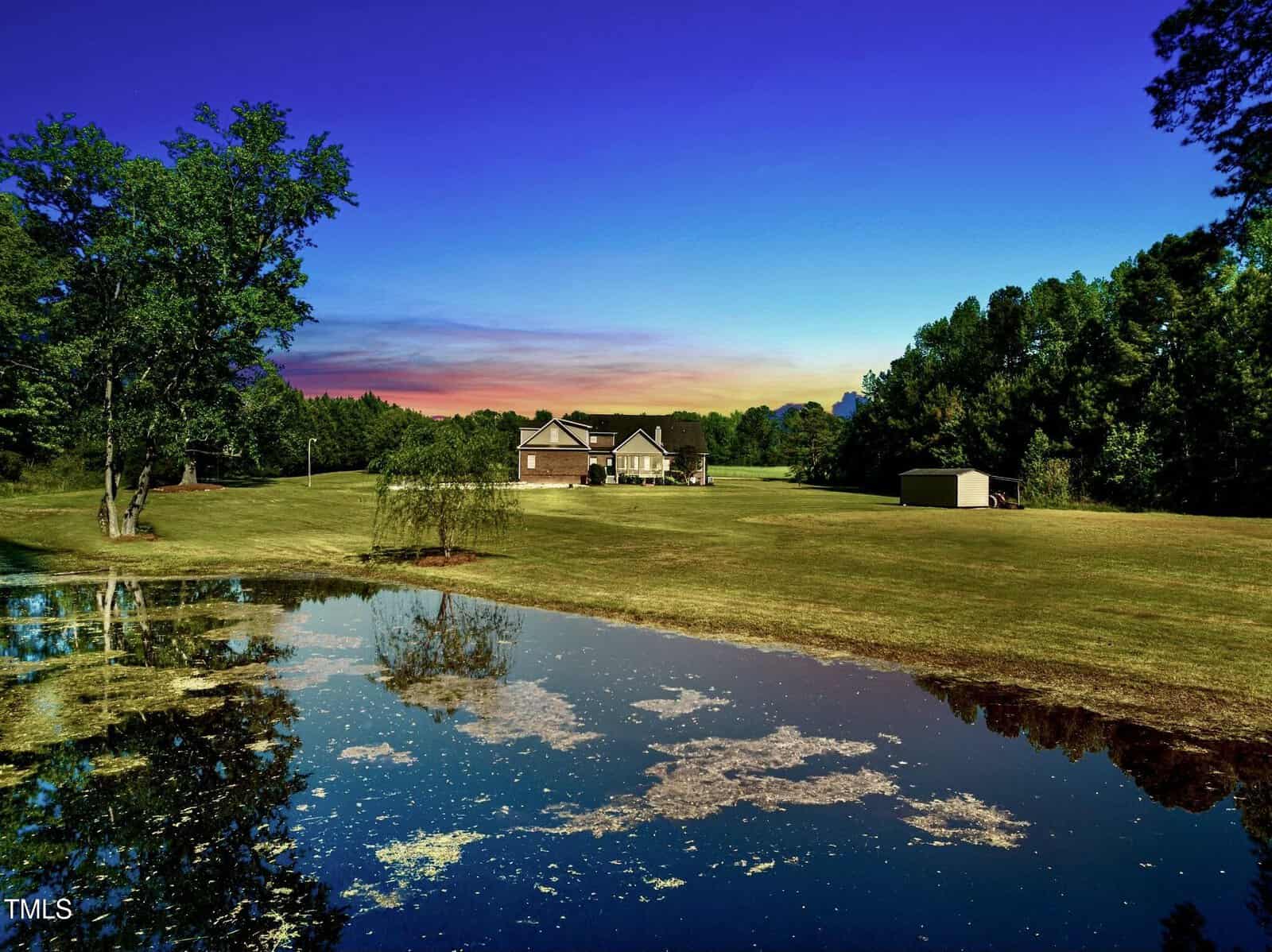 A house with a gray roof sits on a large, grassy lawn surrounded by trees at sunset, with a pond reflecting the sky and landscape in the foreground.