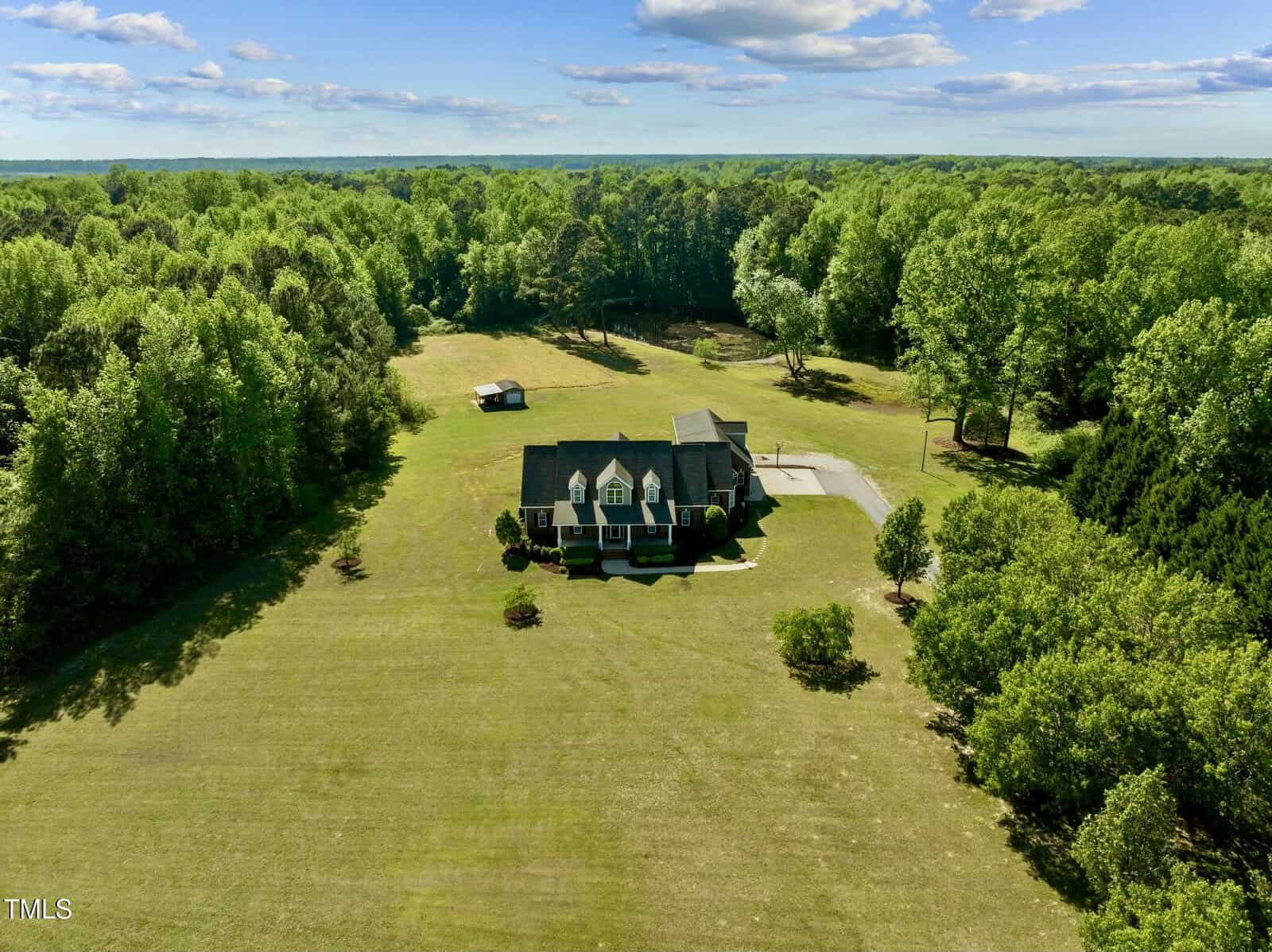 A large house with a dark roof sits in the center of a wide, grassy lawn, surrounded by dense green trees on all sides under a partly cloudy sky. A small outbuilding is visible behind the house.