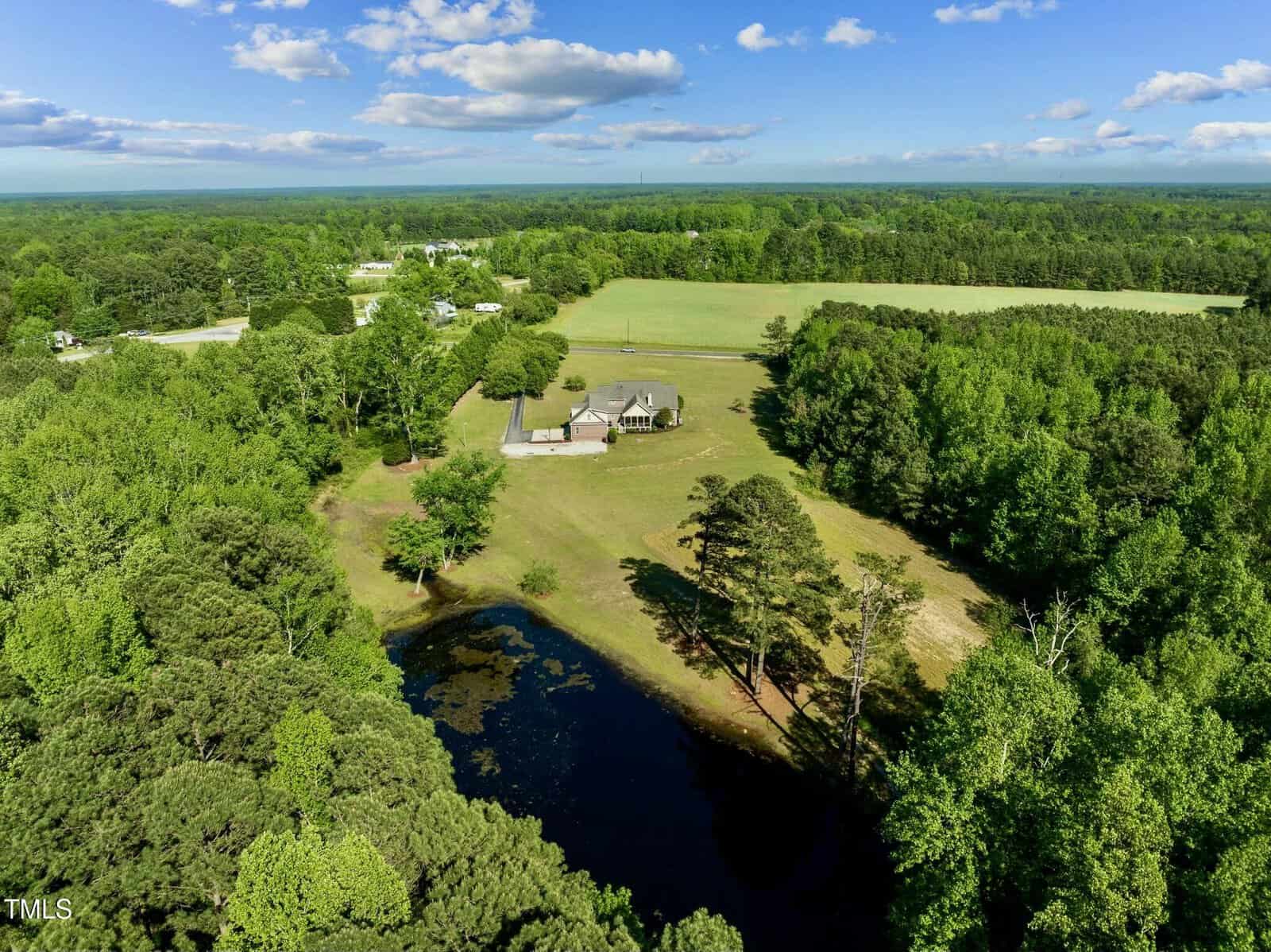 Aerial view of a rural property with a house, open grassy fields, a small pond, and dense trees surrounding the area under a blue sky with scattered clouds.
