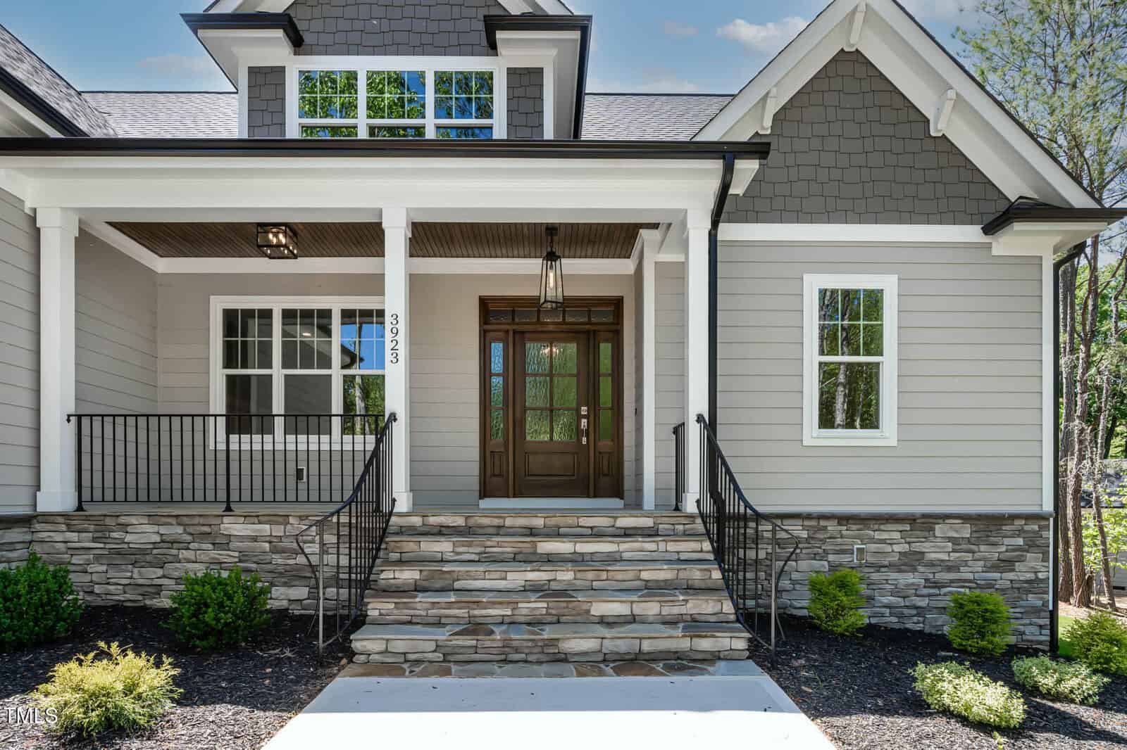 A modern house exterior with gray siding, white trim, a wooden front door with glass panels, two lantern lights, stone steps, metal railings, and small shrubs along a mulch bed.