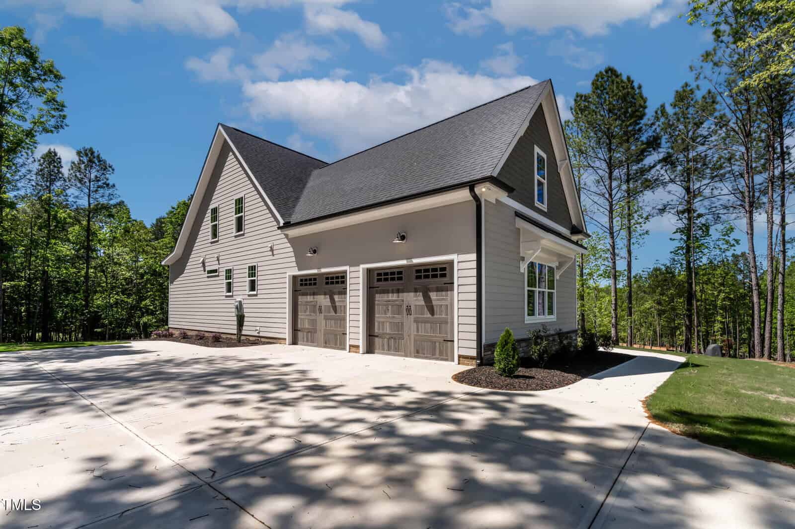 A modern two-story house with light-colored siding, dark roof shingles, and a double garage with wooden doors. The house is surrounded by trees and has a spacious concrete driveway.
