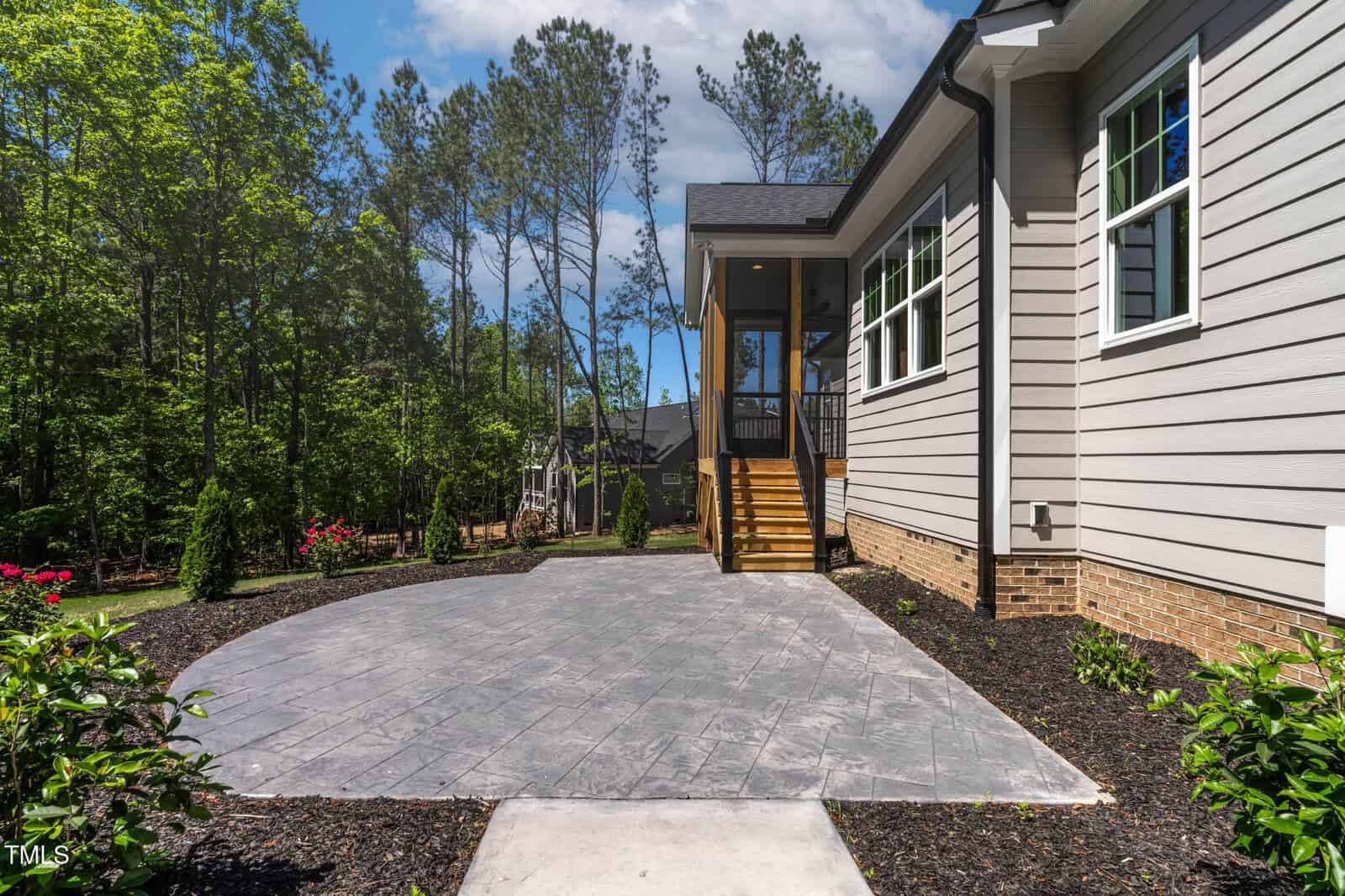 A gray concrete patio with landscaped garden beds and mulch surrounds, next to a house with beige siding and brick foundation. Wooden stairs lead up to a screened porch. Tall trees are visible in the background.