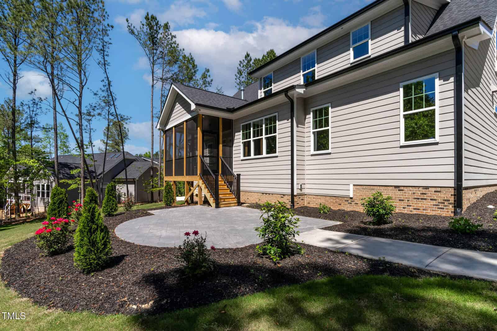 A modern house with gray siding, brick foundation, and a screened porch. The yard features a circular patio, landscaped flower beds, small shrubs, and a concrete walkway. Tall trees and other houses are visible in the background.
