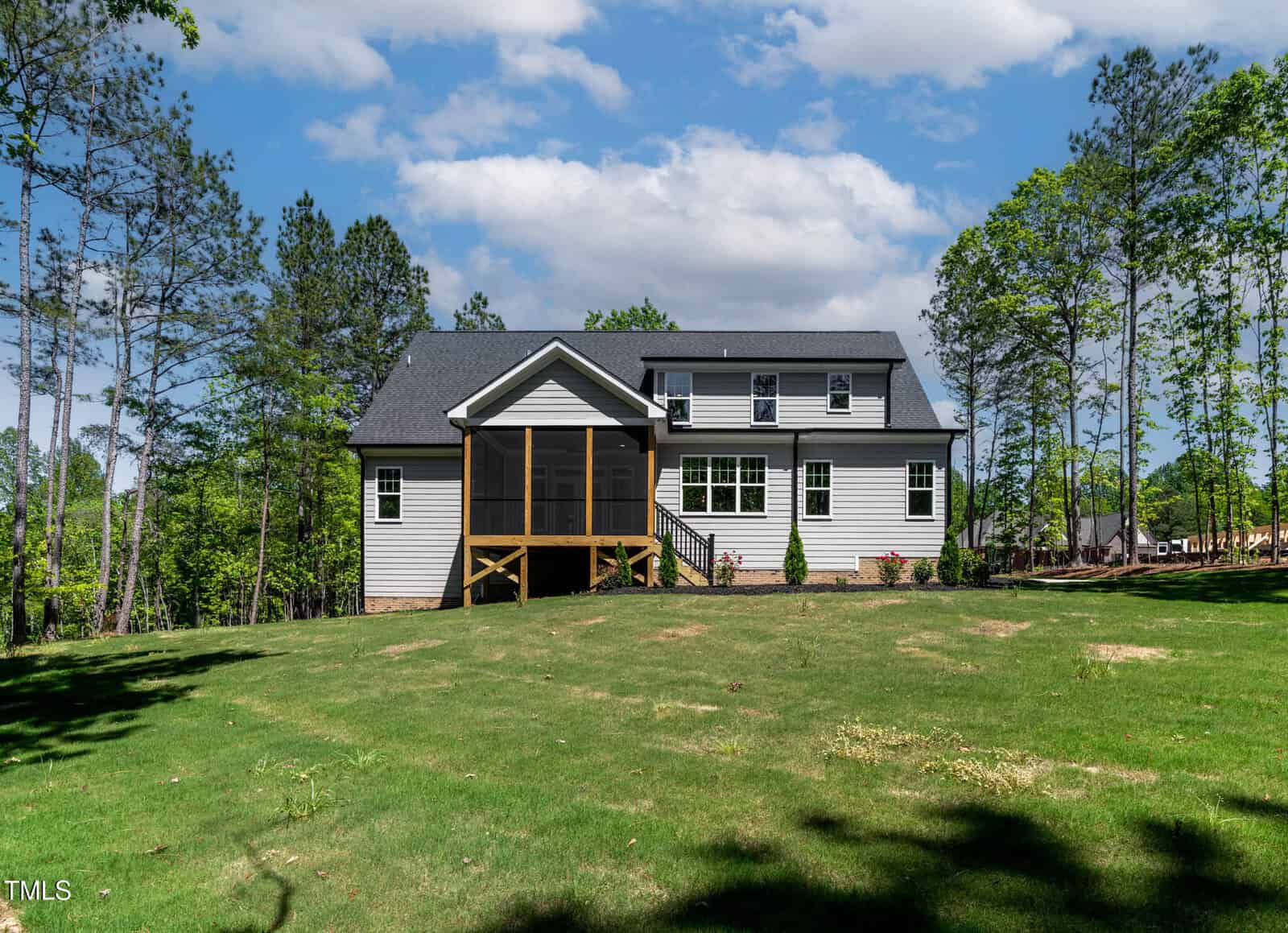 A two-story house with light-colored siding and a screened porch sits on a large, grassy yard, surrounded by tall trees under a partly cloudy sky.