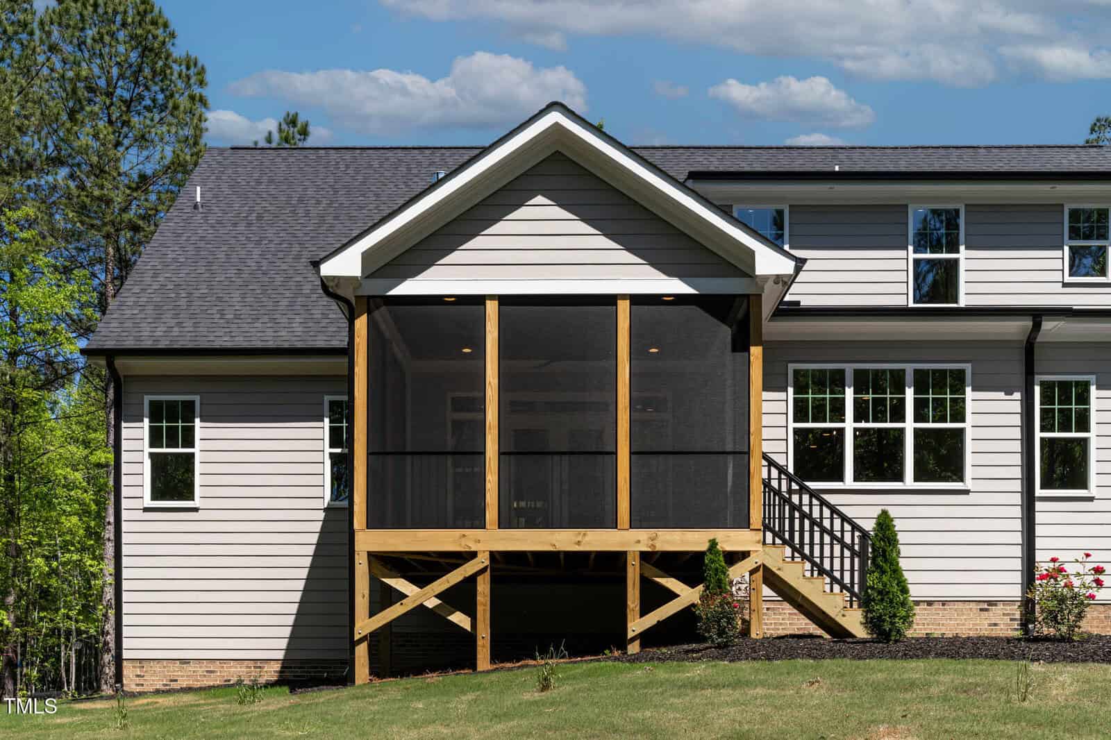 A beige house with white trim features a screened-in porch elevated on wooden posts, accessible by stairs. The lawn is grassy, and several windows are visible along the side of the house. Trees and blue sky are in the background.