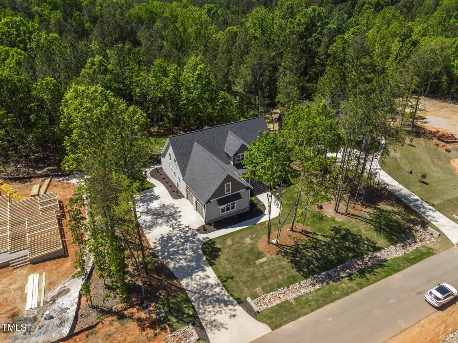 Aerial view of a gray two-story house with a dark roof, surrounded by trees and green lawn, situated on a spacious lot with a paved driveway in a suburban neighborhood under construction.
