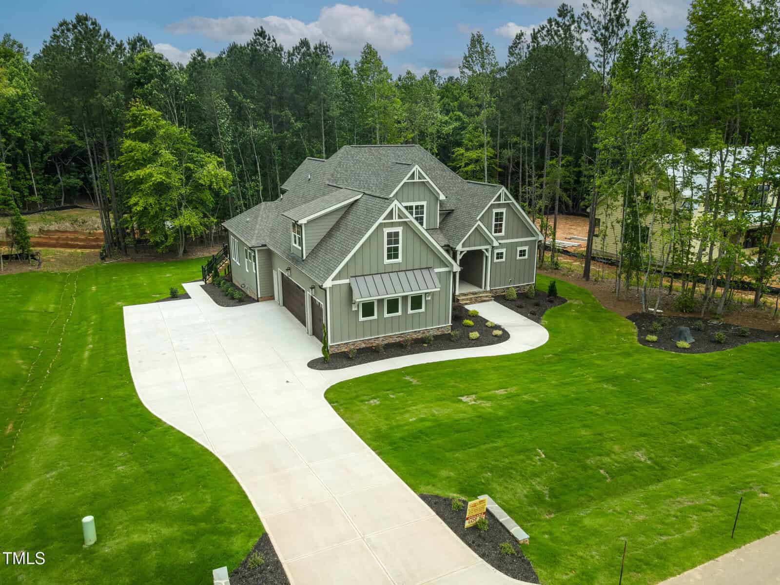 A large two-story house with gray siding and a three-car garage sits on a spacious, neatly landscaped lawn with a curved concrete driveway, surrounded by trees and greenery.